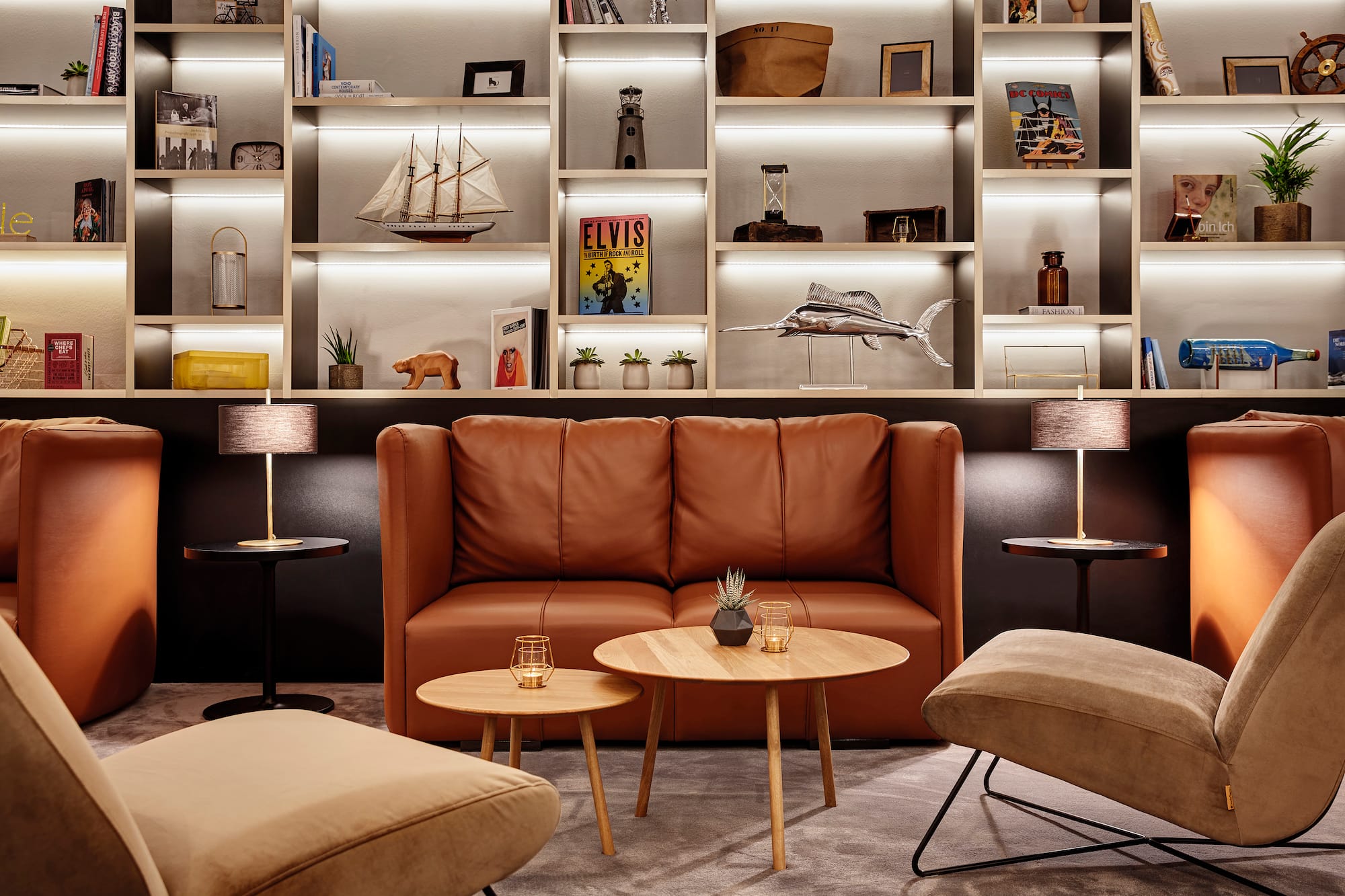 Brown leather sofa, accent chairs, and illuminated bookshelf in modern lounge.