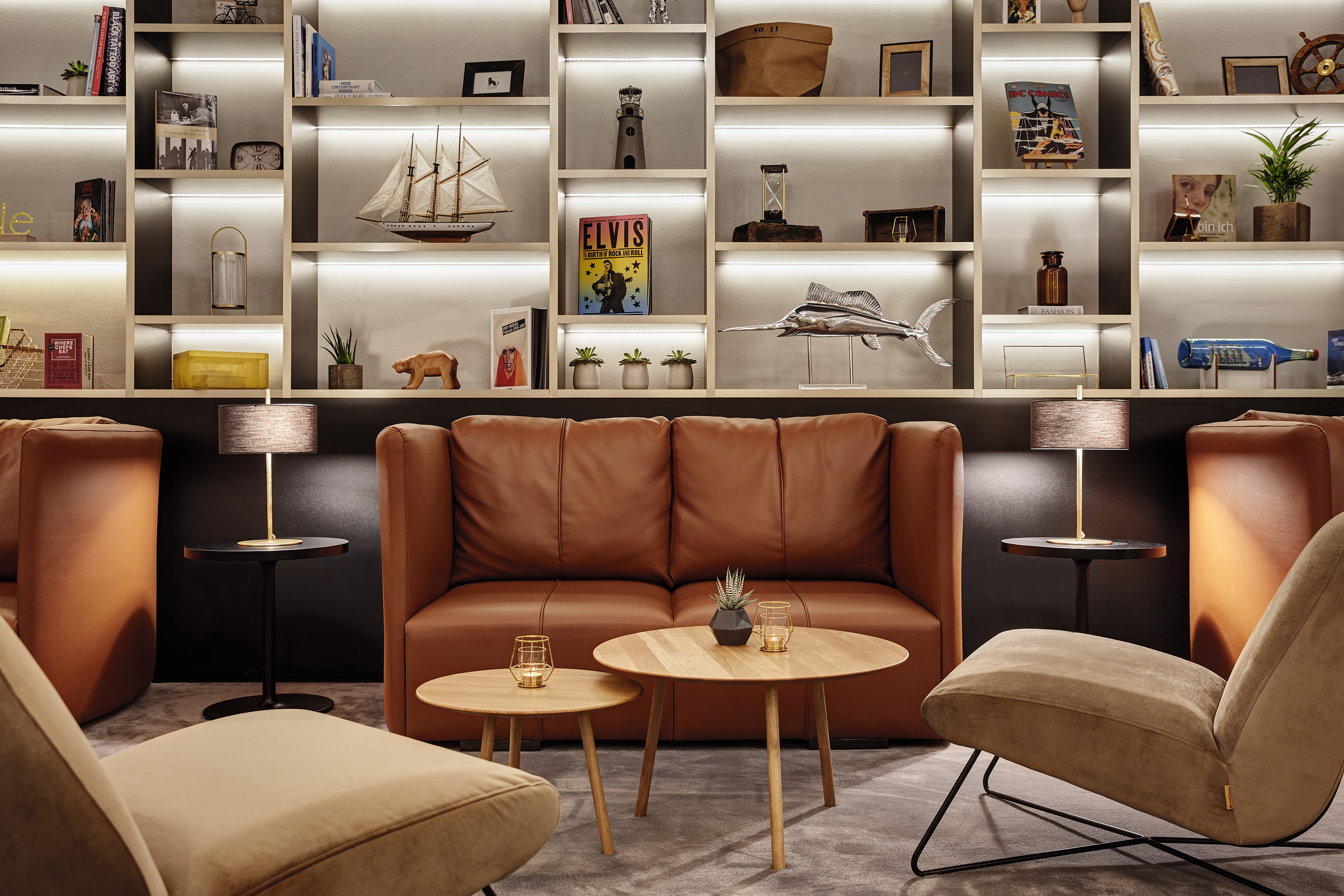 Brown leather sofa, accent chairs, and illuminated bookshelf in modern lounge.