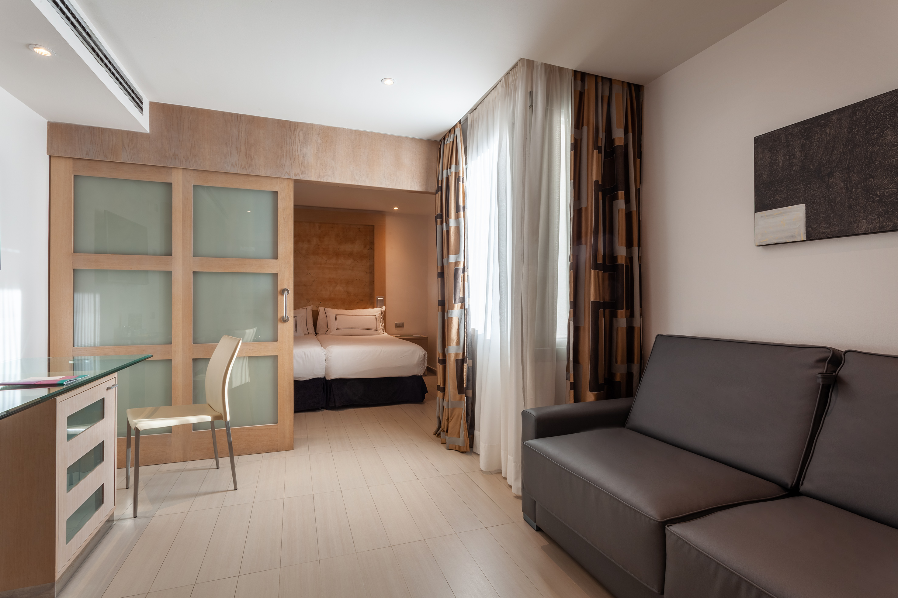 Contemporary hotel studio with sofa, desk, and bedroom behind sliding doors.