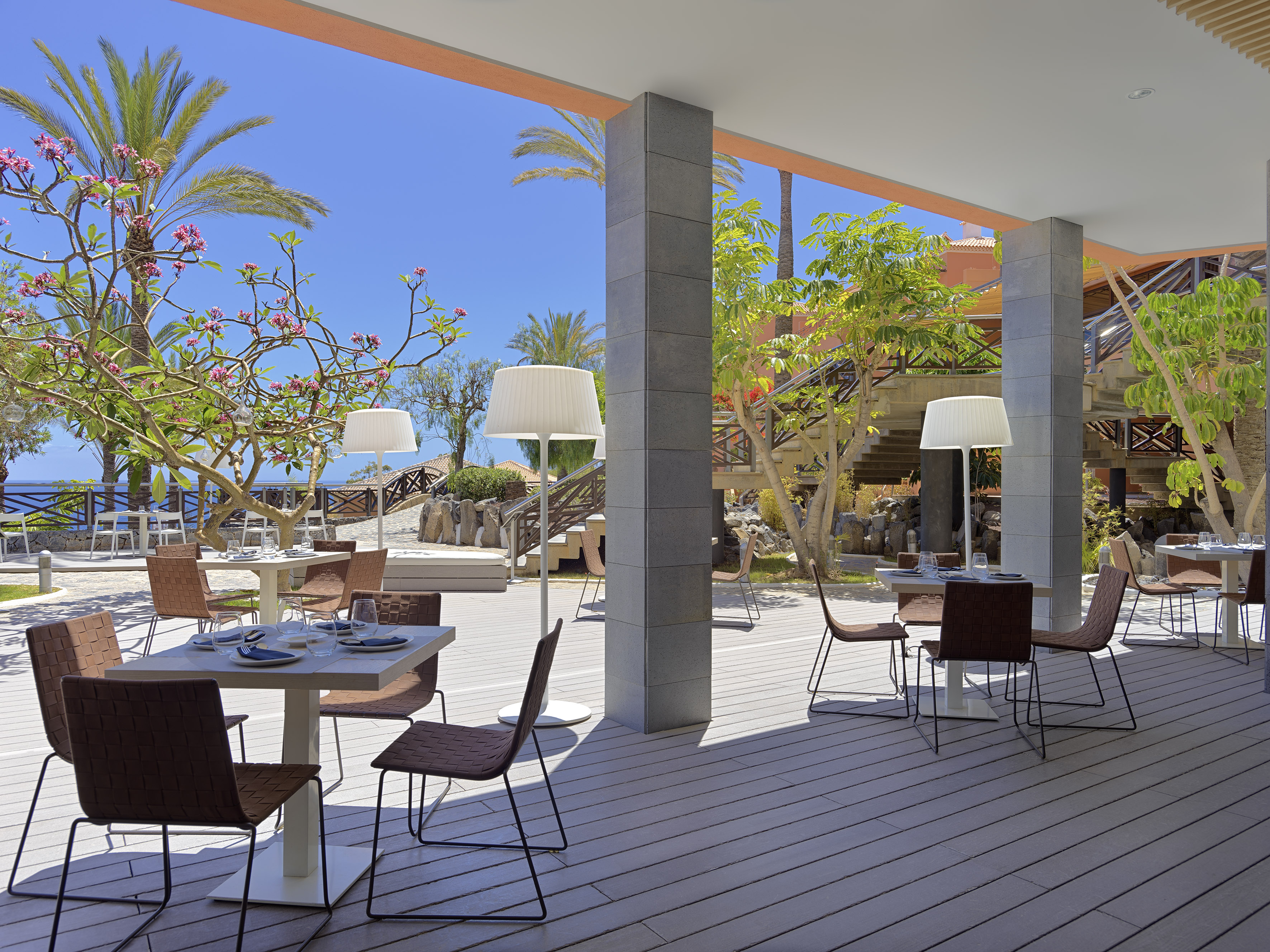 Resort al fresco dining area with tables, chairs, palm trees, and sea view.