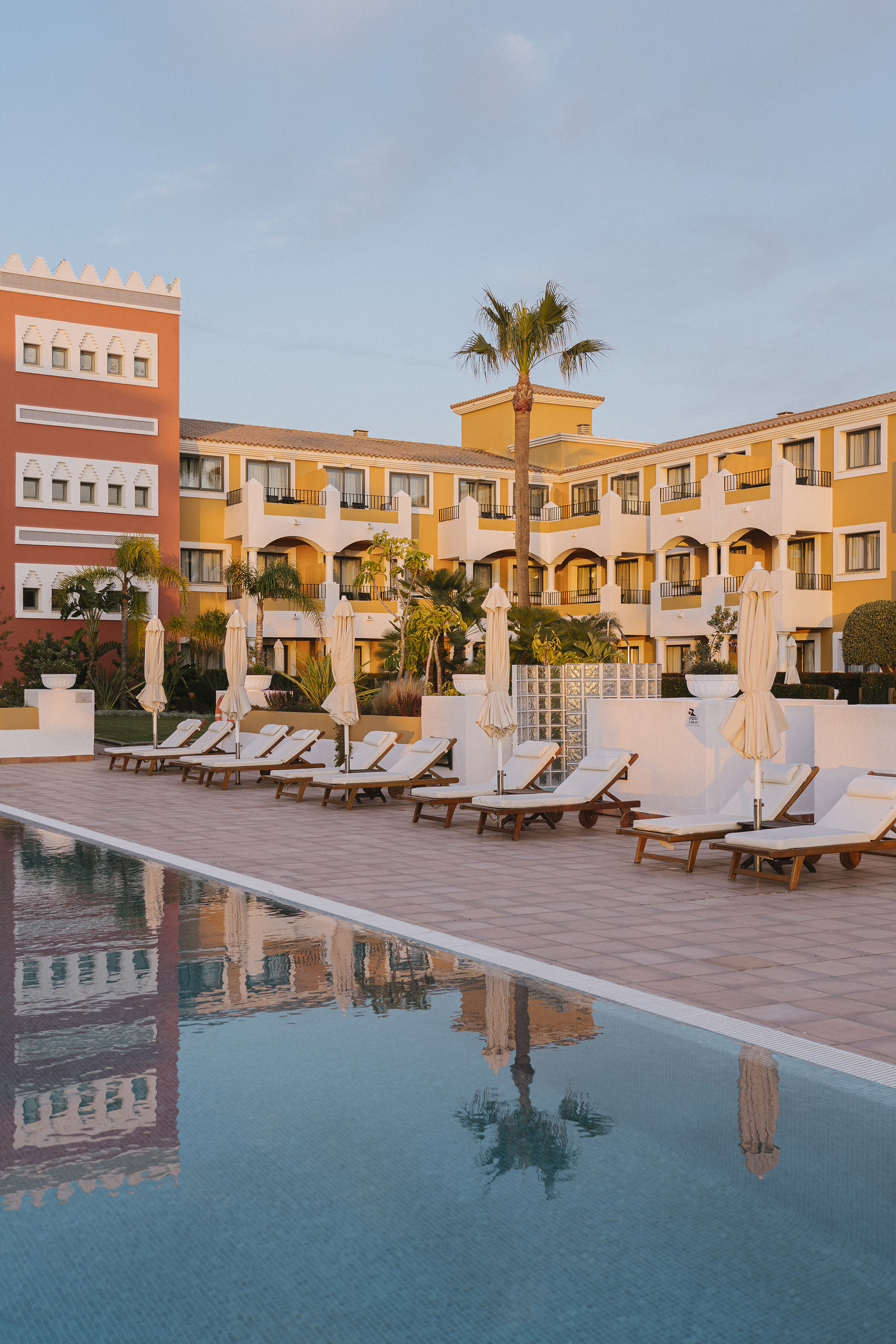 Resort pool with sun loungers, palm trees, and yellow/red buildings at sunset.