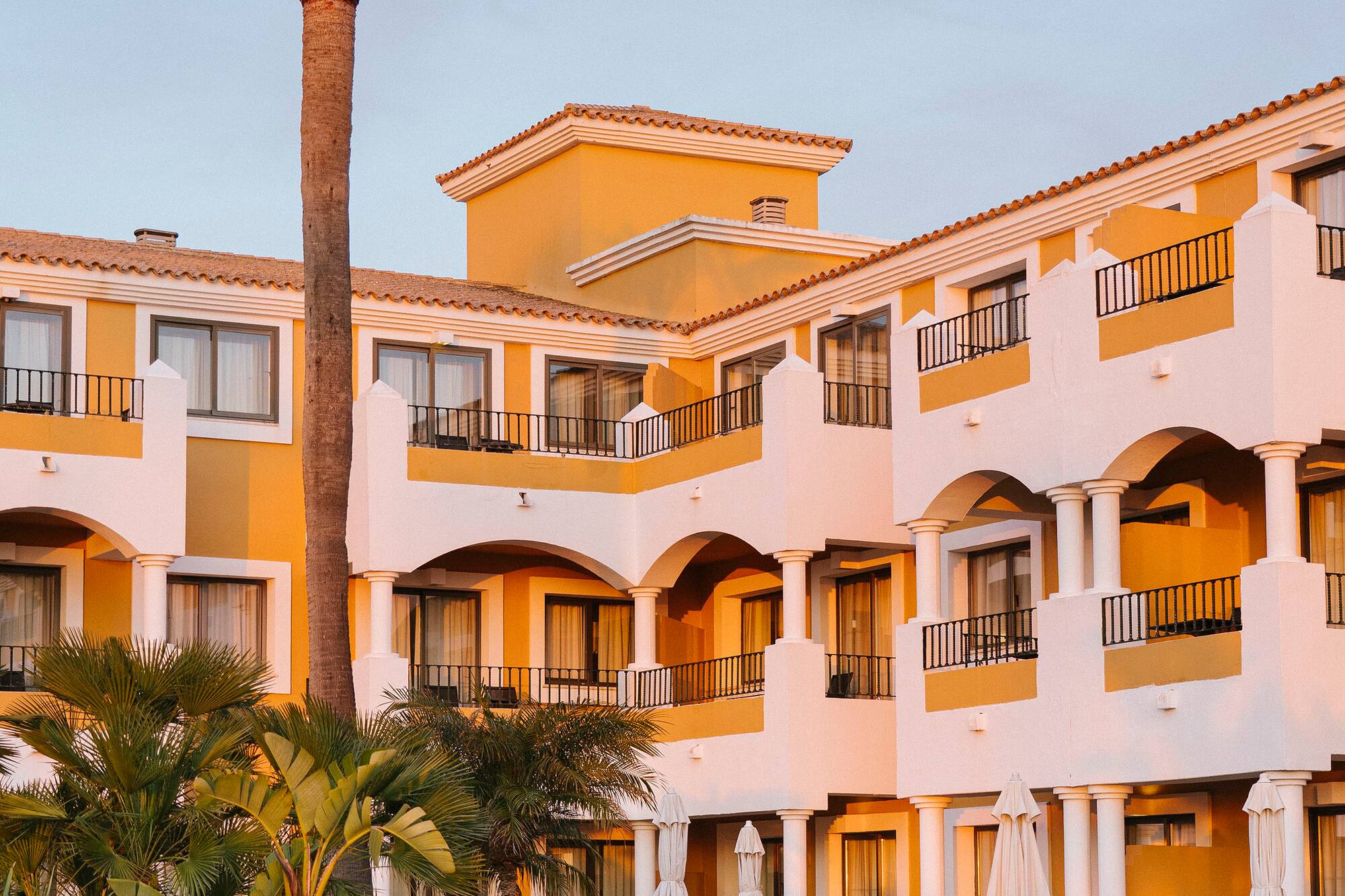 Mediterranean resort with yellow walls, balconies, palm trees, and garden.