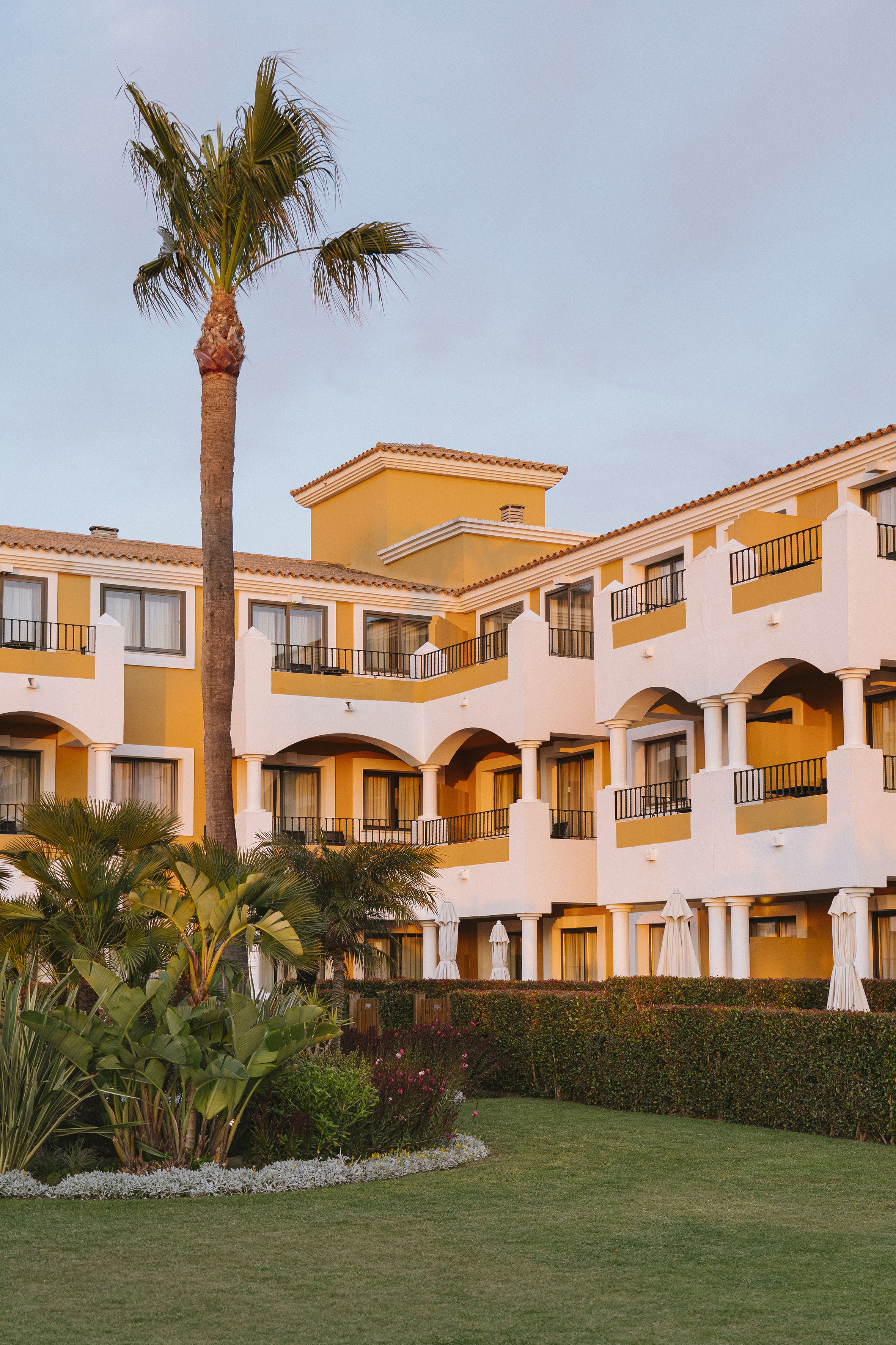 Mediterranean resort with yellow walls, balconies, palm trees, and garden.