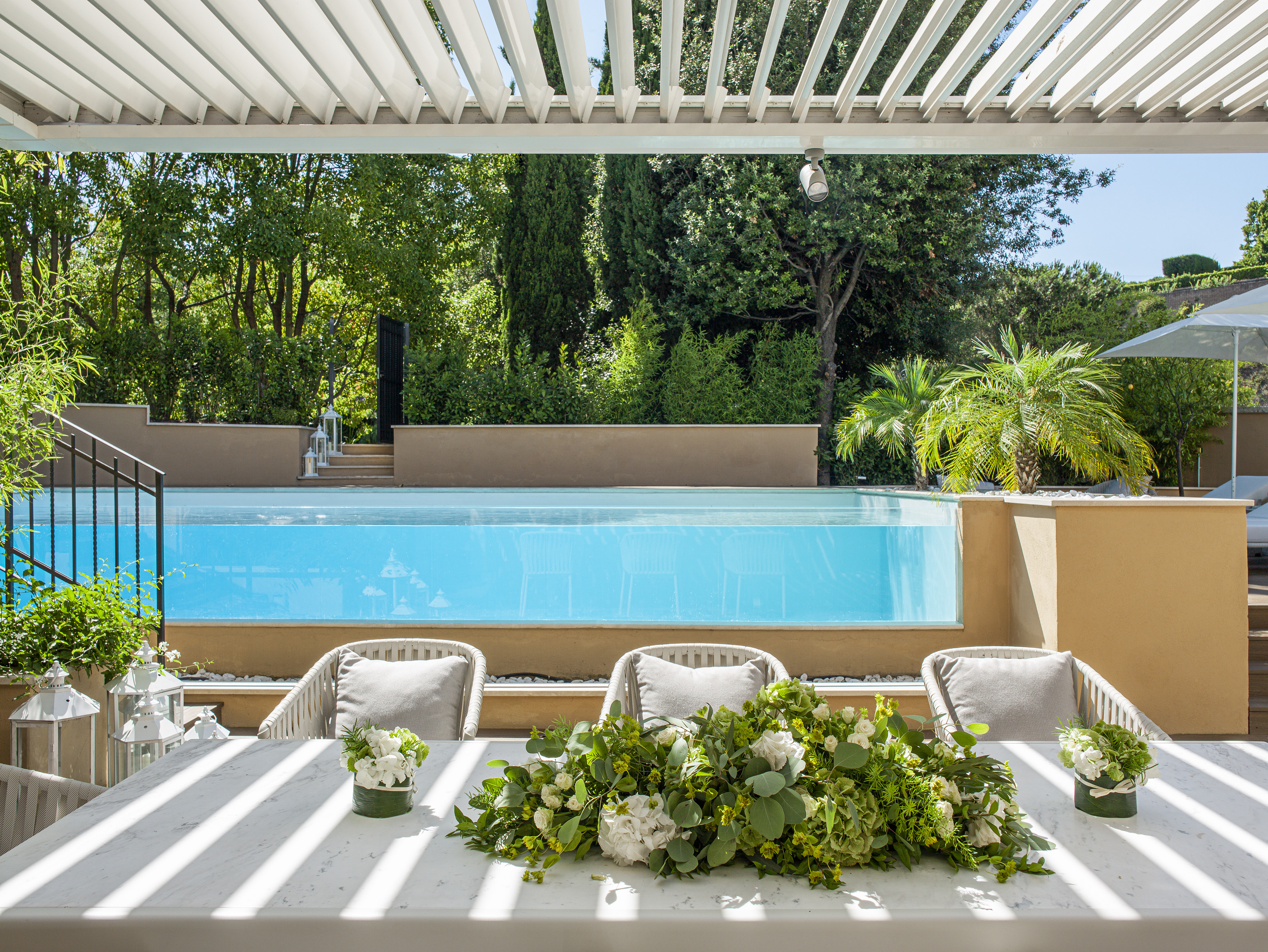 Luxury outdoor dining patio with pergola, clear pool, and lush garden.