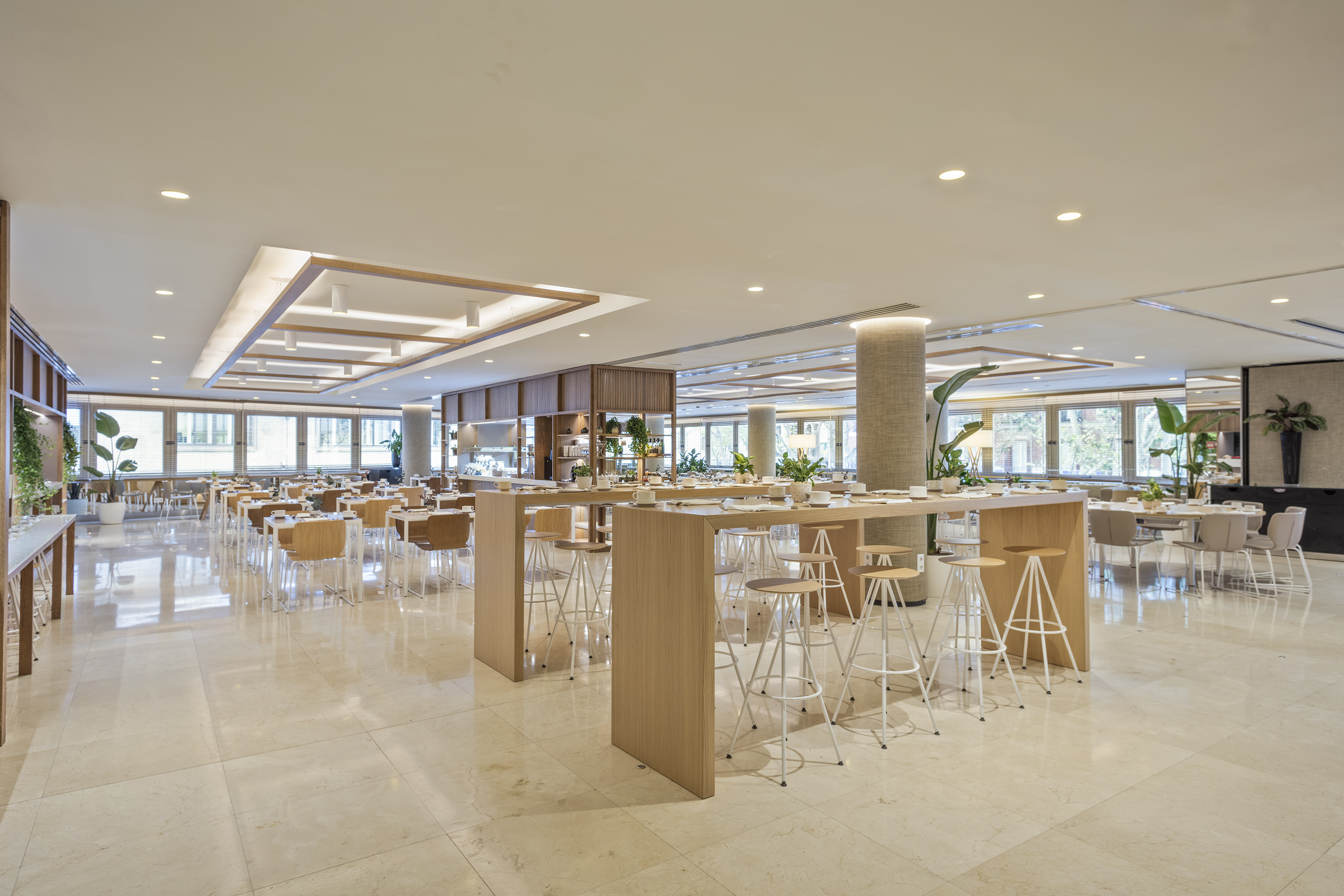Modern hotel dining room with light wood tables, bar stools, and marble floor.