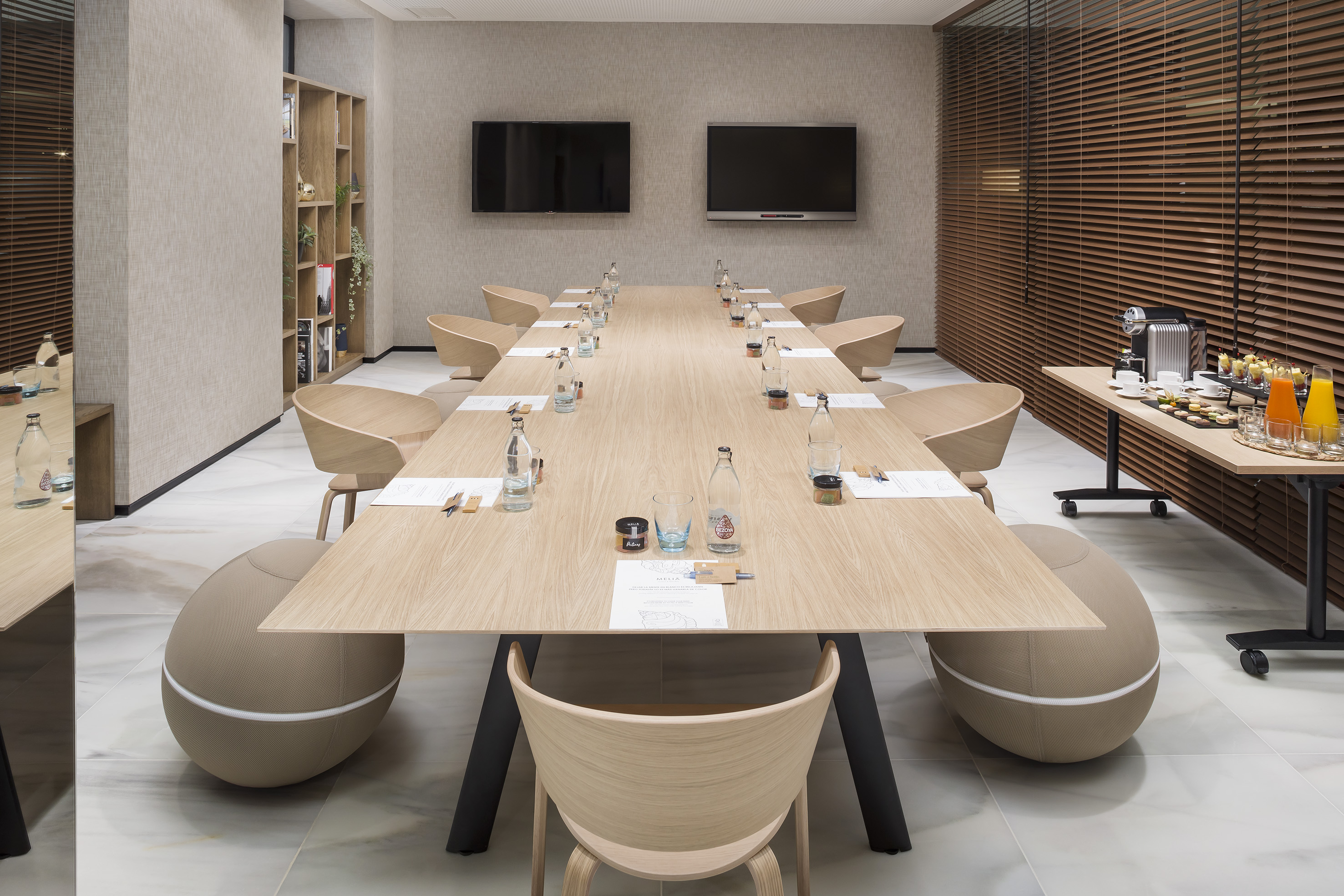 Modern meeting room with long wooden table, screens, and refreshment station.