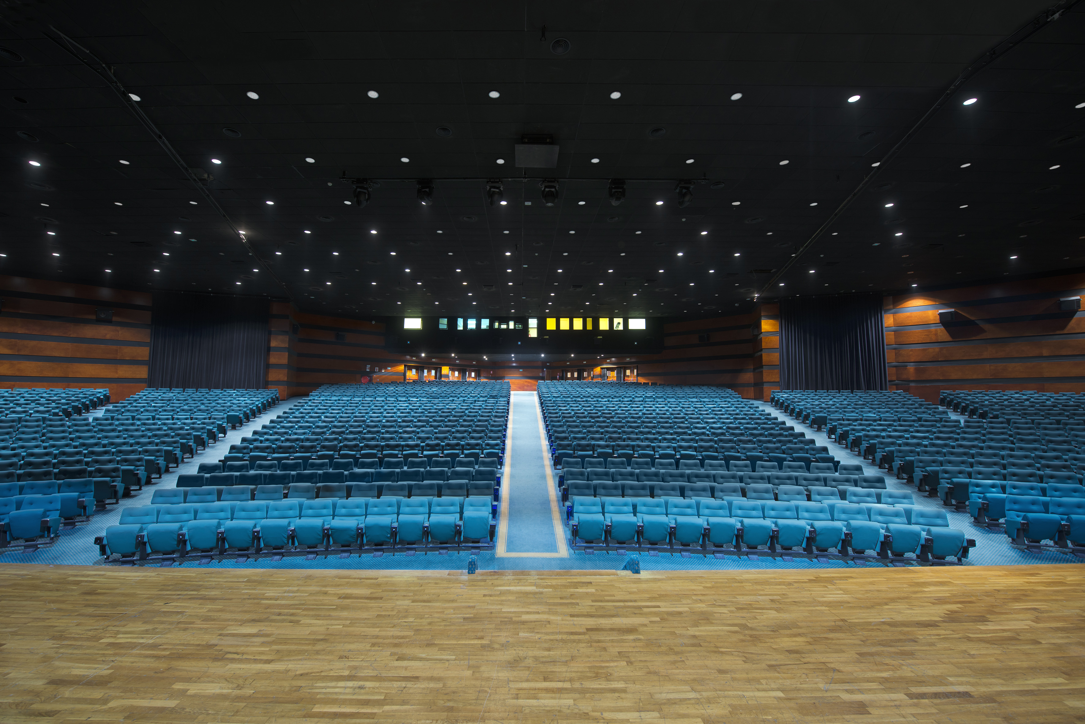 Large empty auditorium with blue tiered seating and wooden stage.