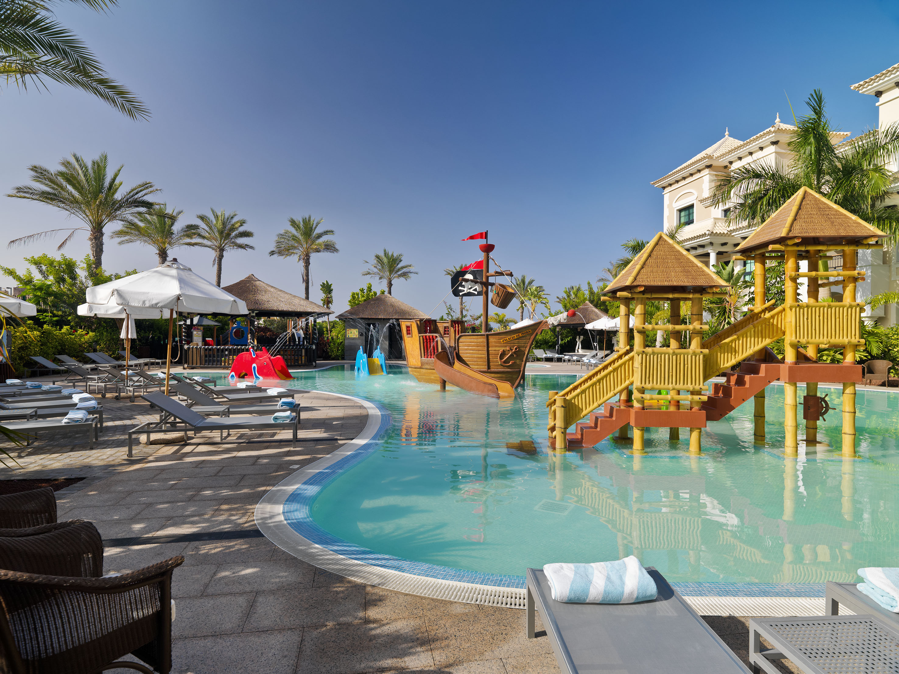Sunny family resort pool with pirate ship play area, palm trees, and lounge chairs.