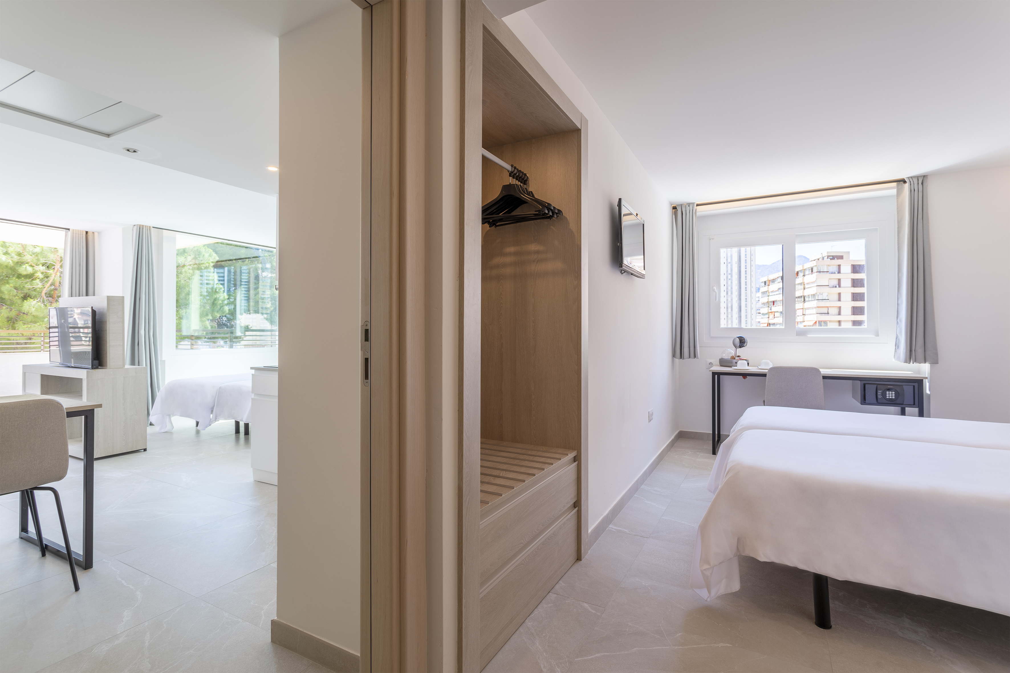 Modern hotel twin bedroom with desk, wardrobe, and a bright adjoining room.