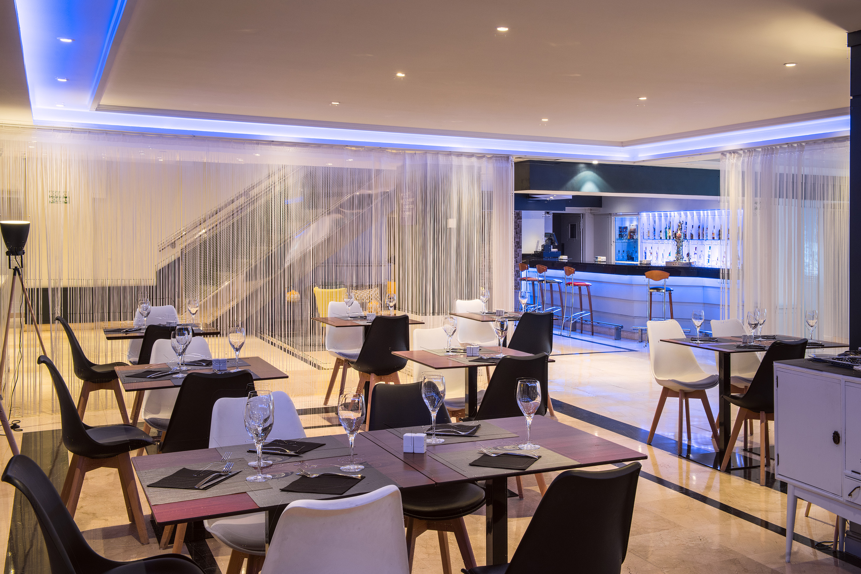 Modern restaurant dining room with set tables, bar, and blue accent lighting.