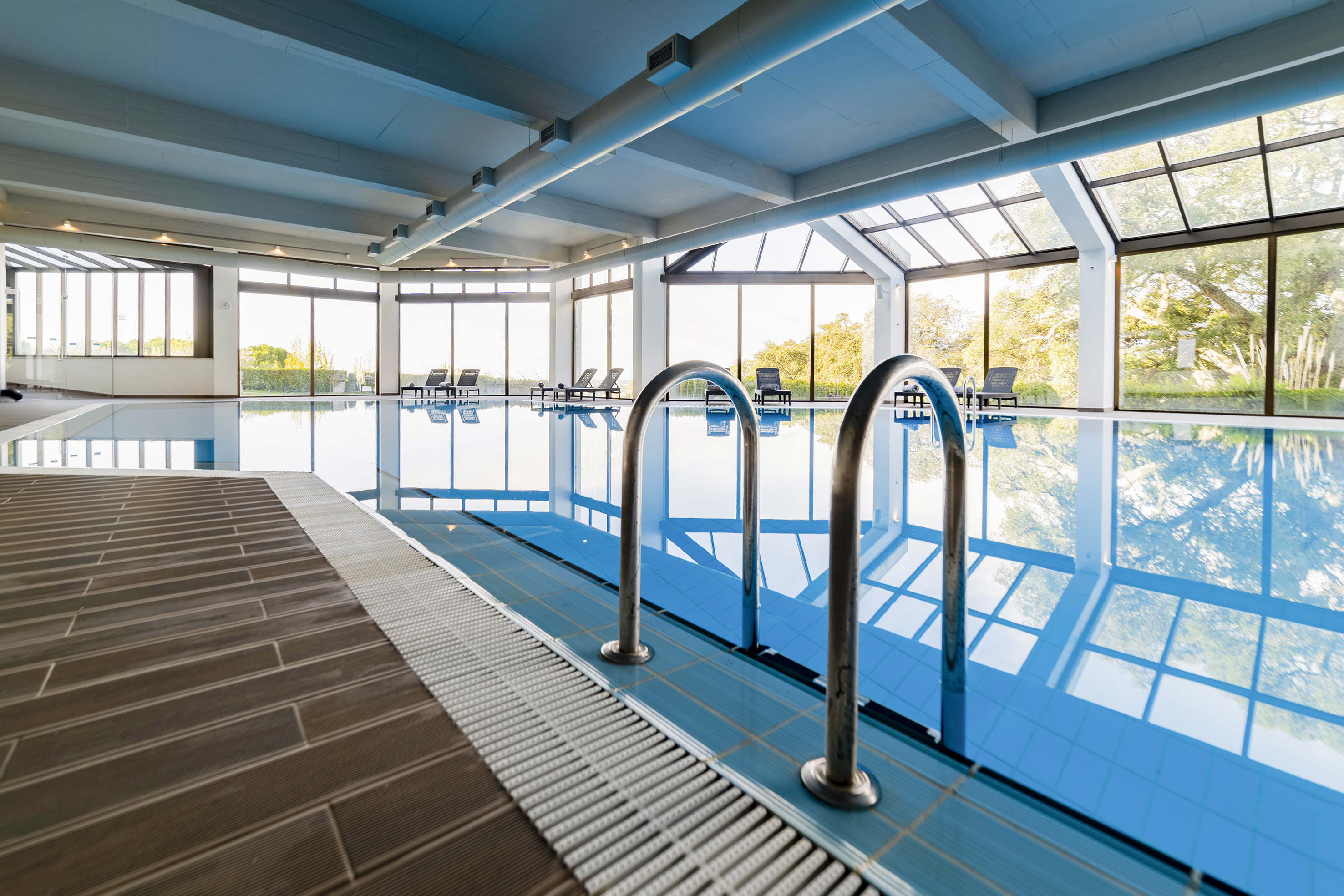Modern indoor swimming pool with blue water, large windows, and lounge chairs.