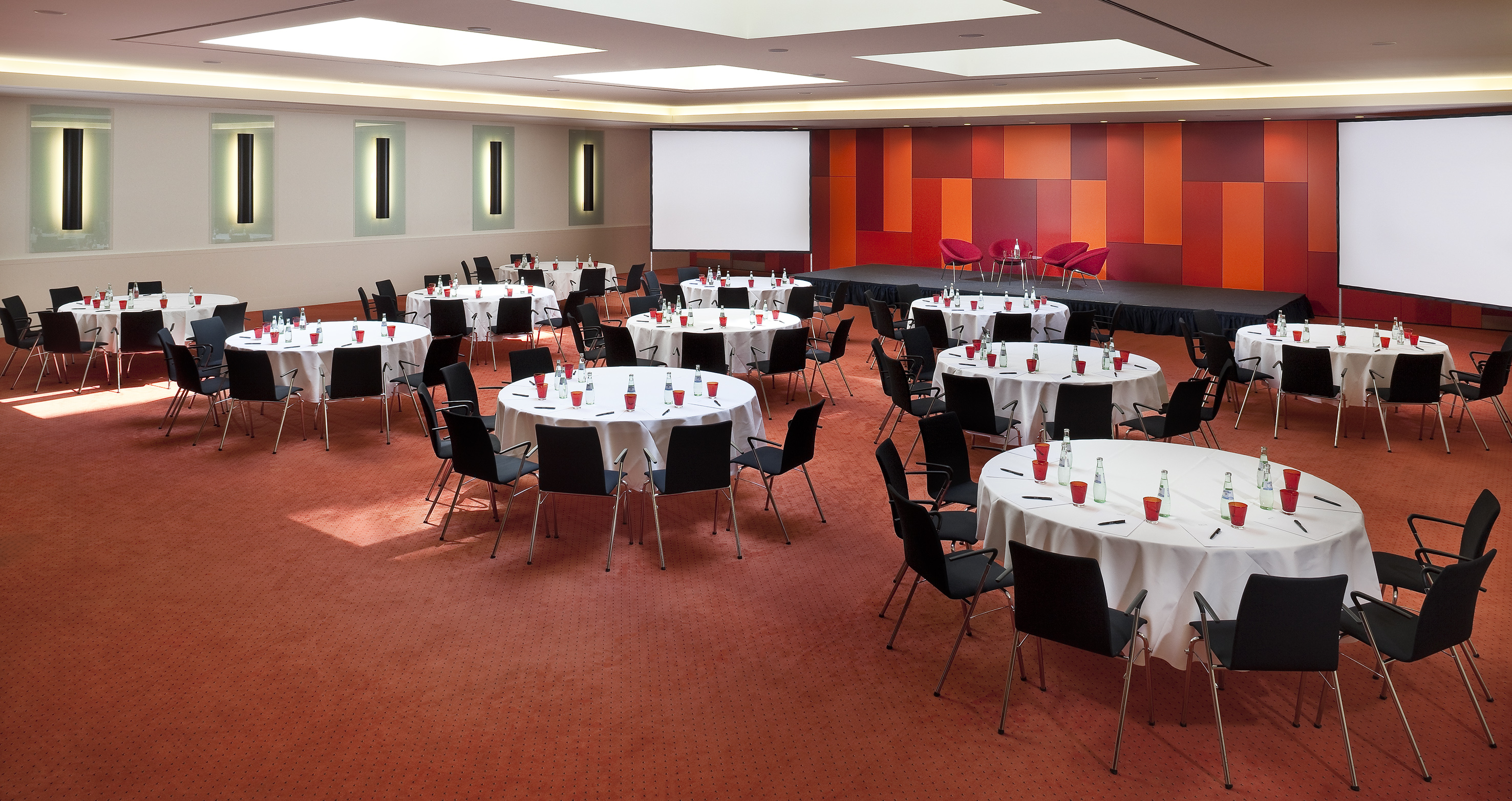 Conference room setup with round tables, chairs, red carpet, stage, and screens.