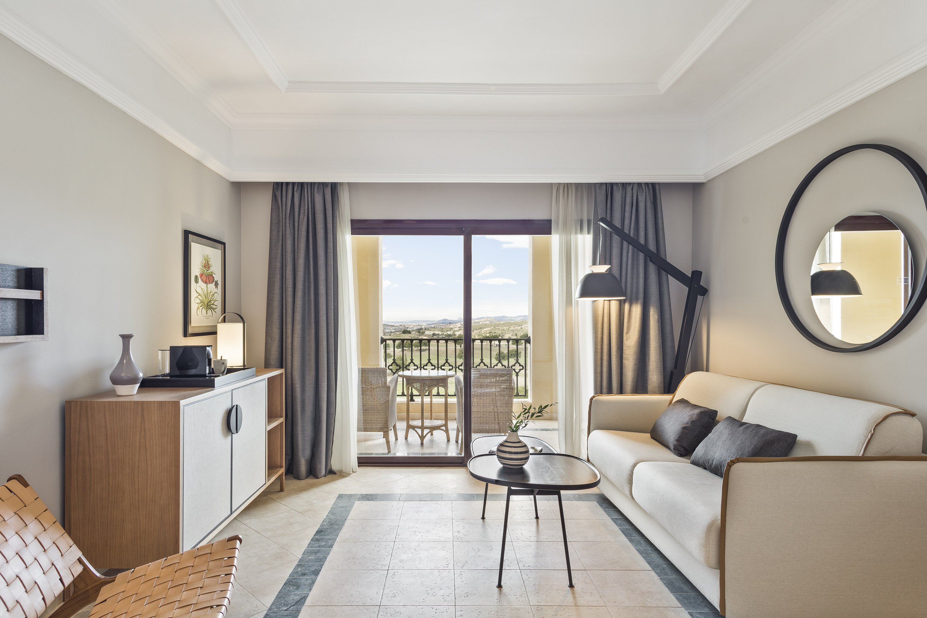 Modern hotel suite living room with sofa, balcony, and scenic landscape view.