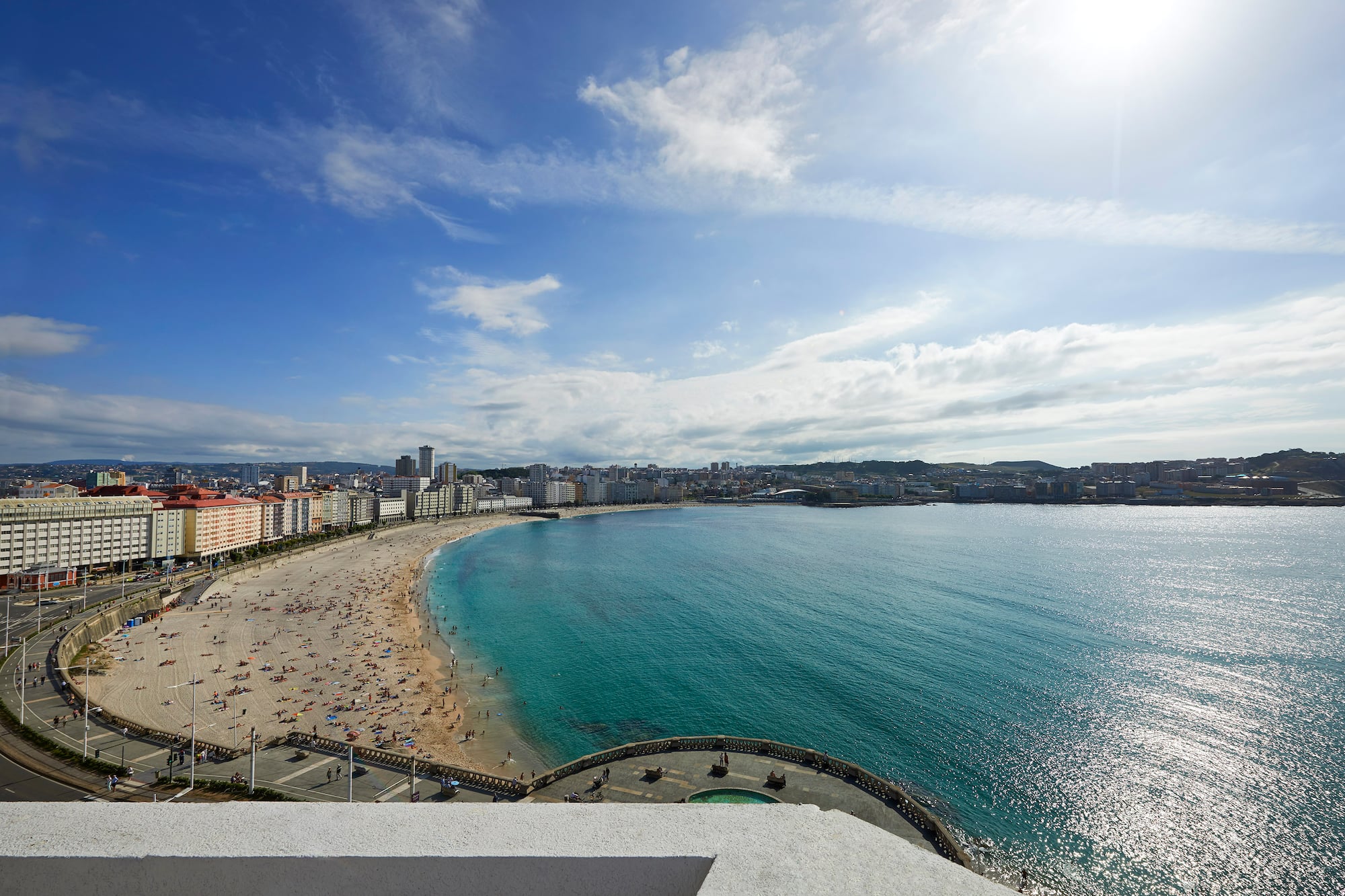 Crowded city beach, turquoise ocean, and coastal city under a sunny sky.