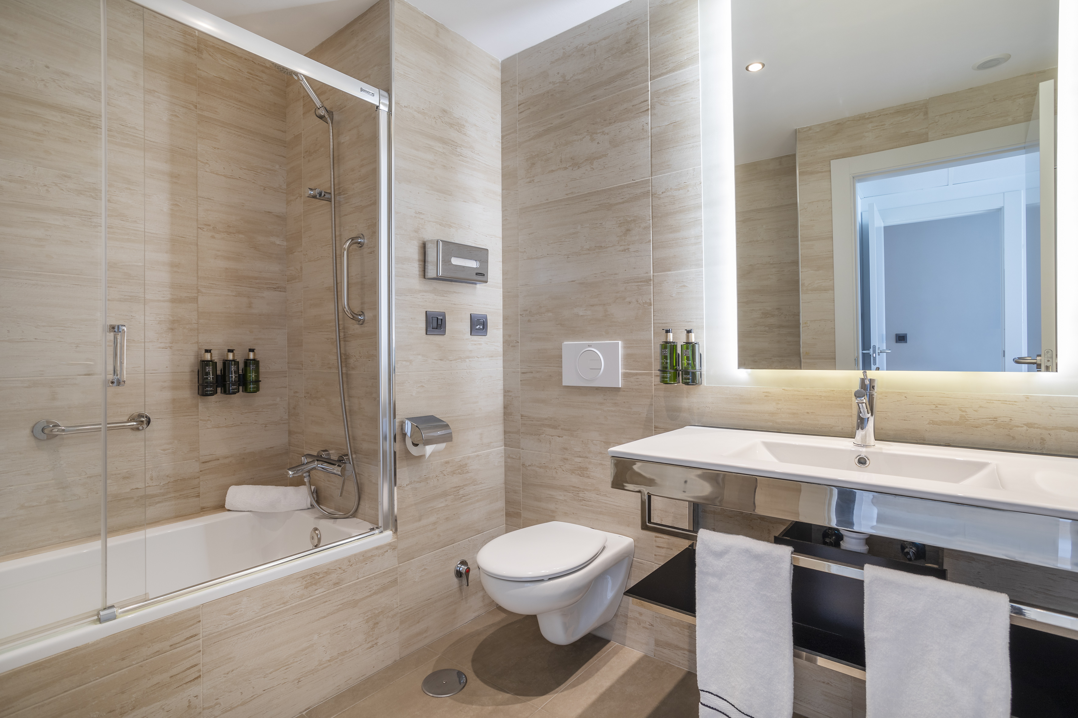 Modern hotel bathroom with shower bath, wall-mounted toilet, and backlit vanity mirror.