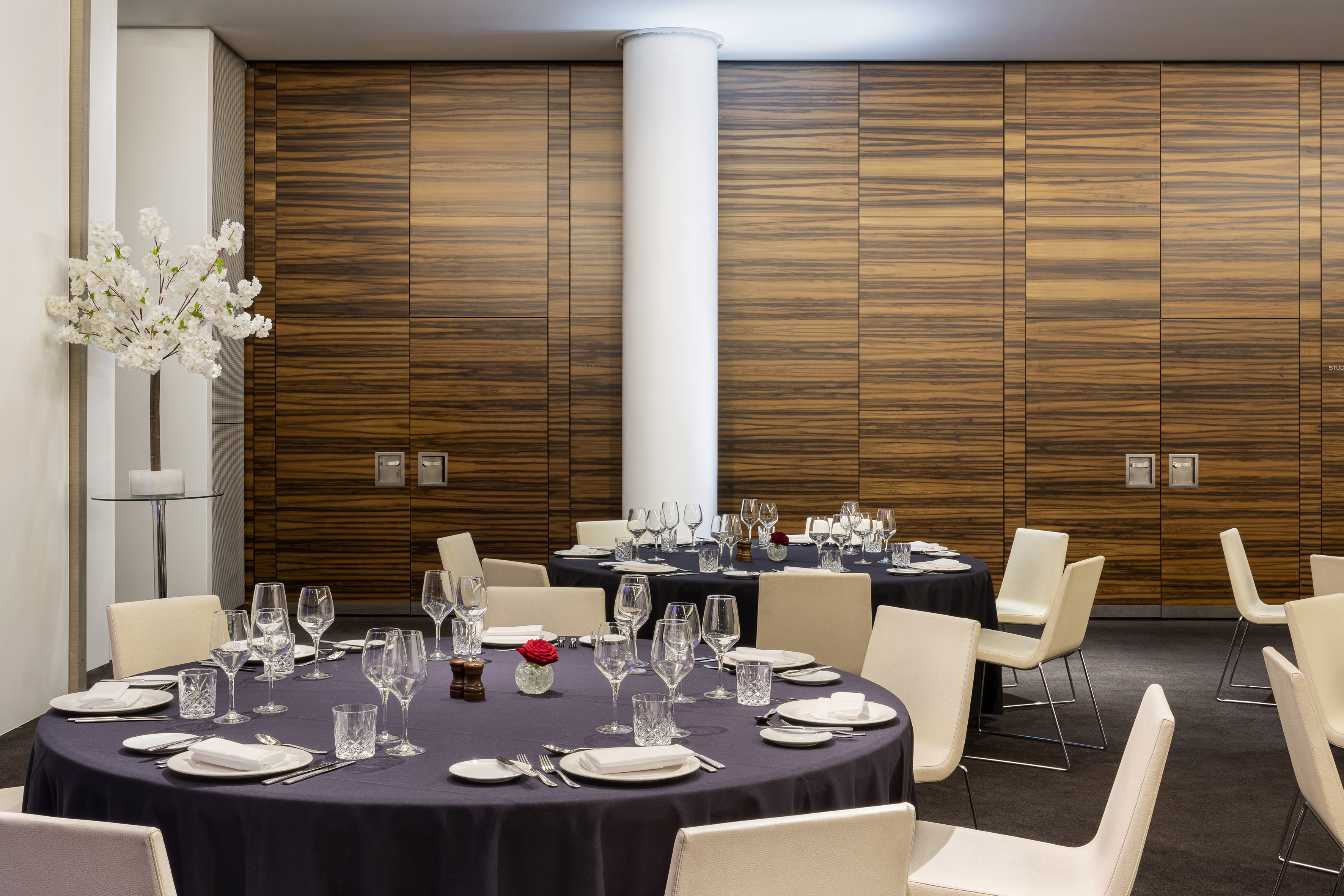 Elegant event space with set dining tables, wood-paneled walls, and cream chairs.