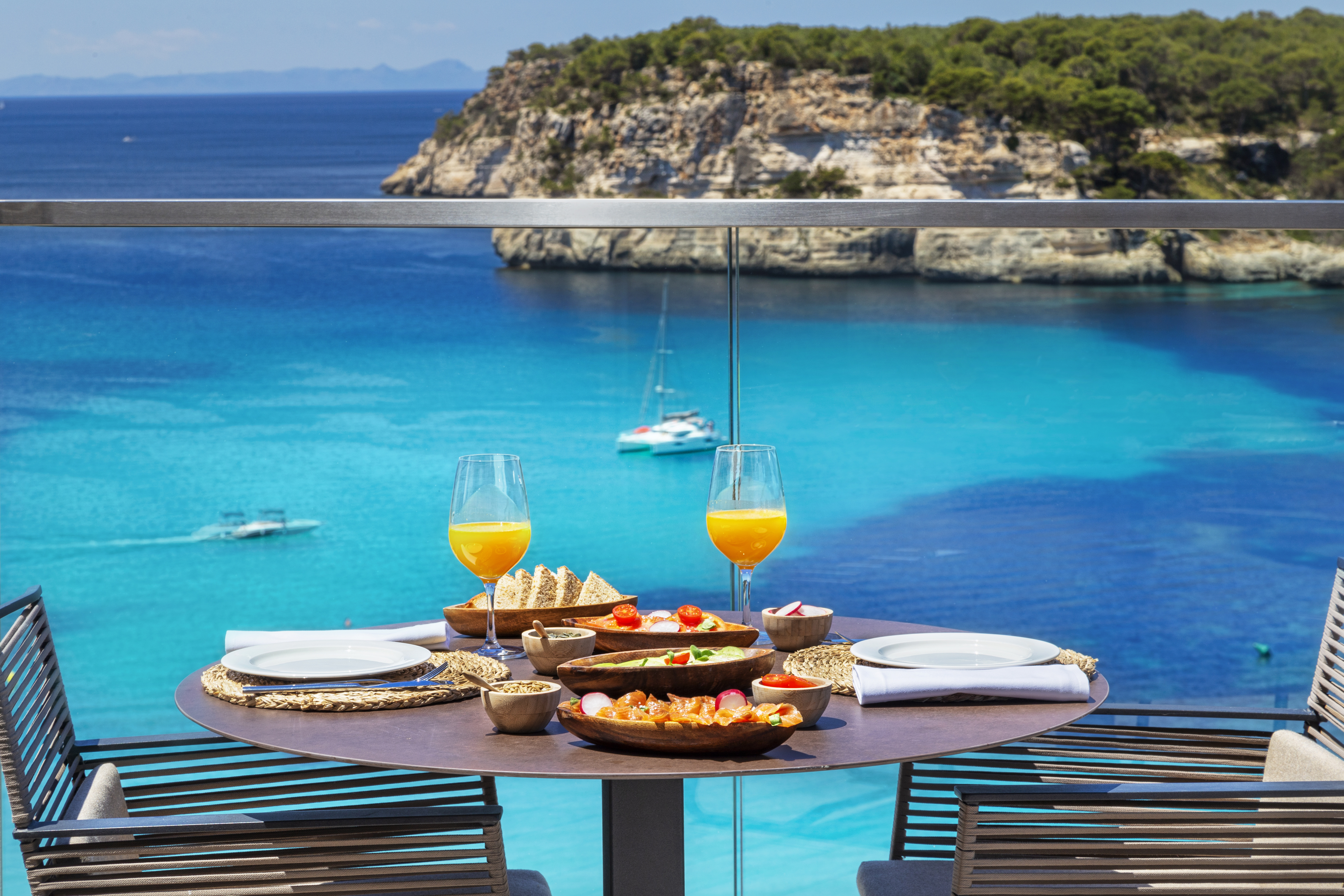 Balcony brunch with vibrant blue ocean, sailboat, and rocky coastline.