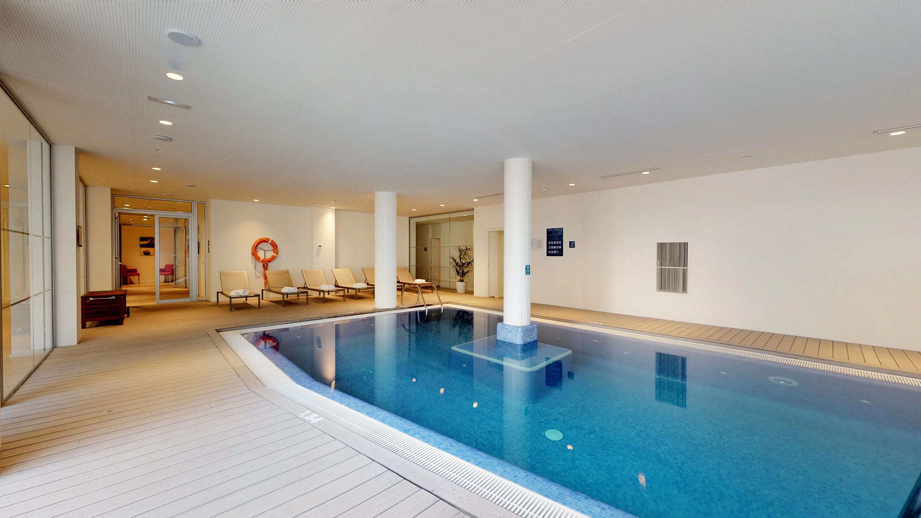 Modern indoor swimming pool with blue water, lounge chairs, and white walls.