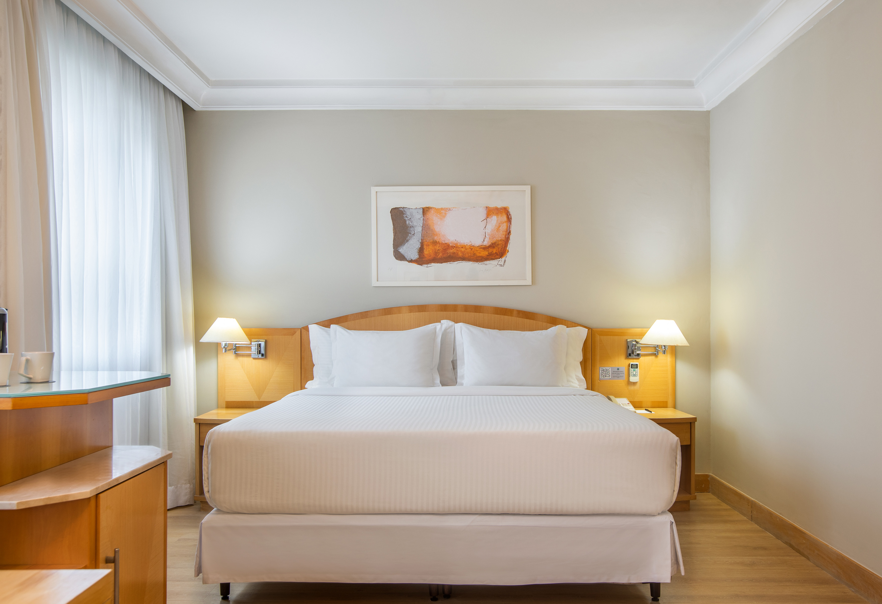 Modern hotel room with king bed, wood headboard, lamps, and abstract artwork.
