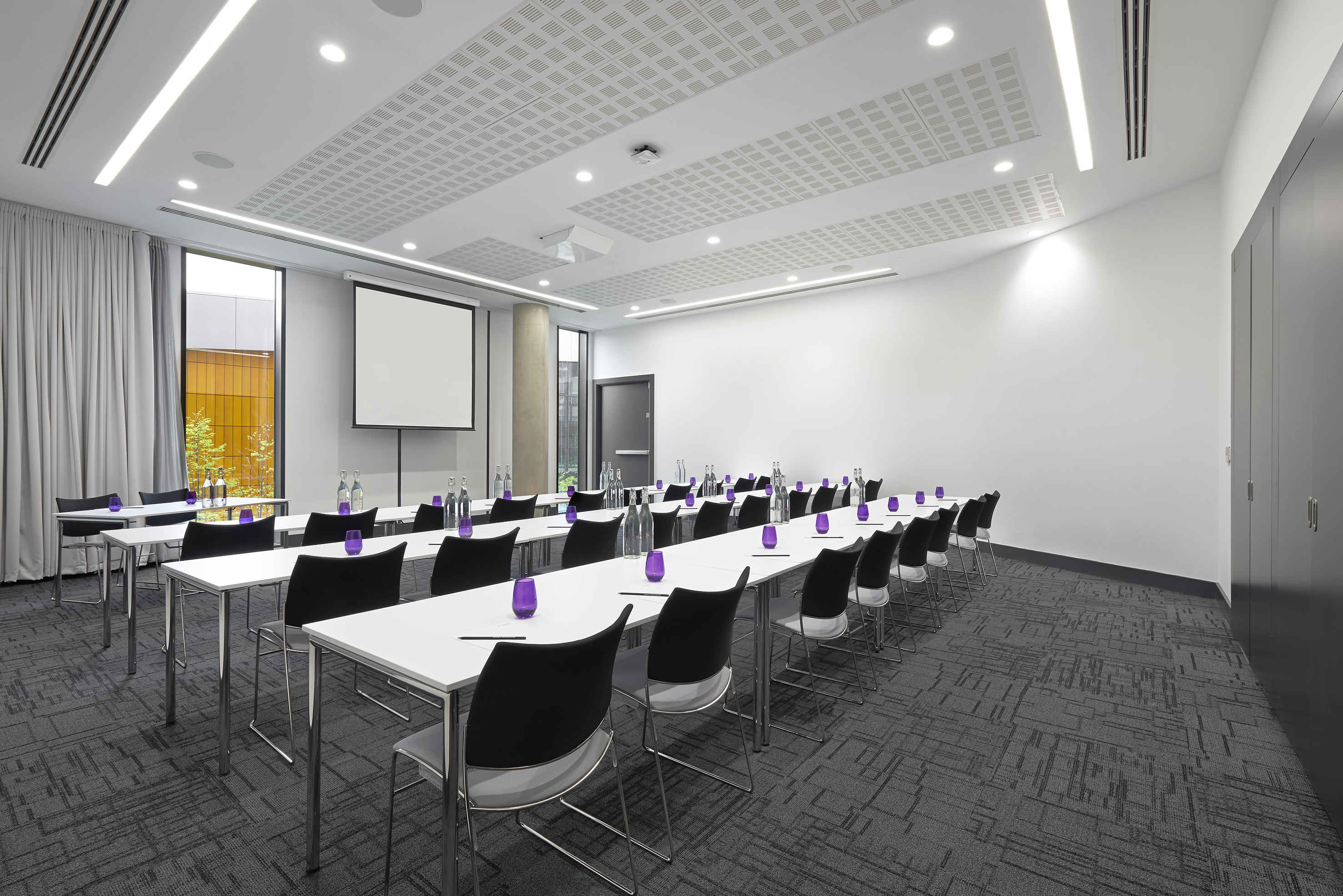 Modern conference room with tables, chairs, projector, screen, natural light.