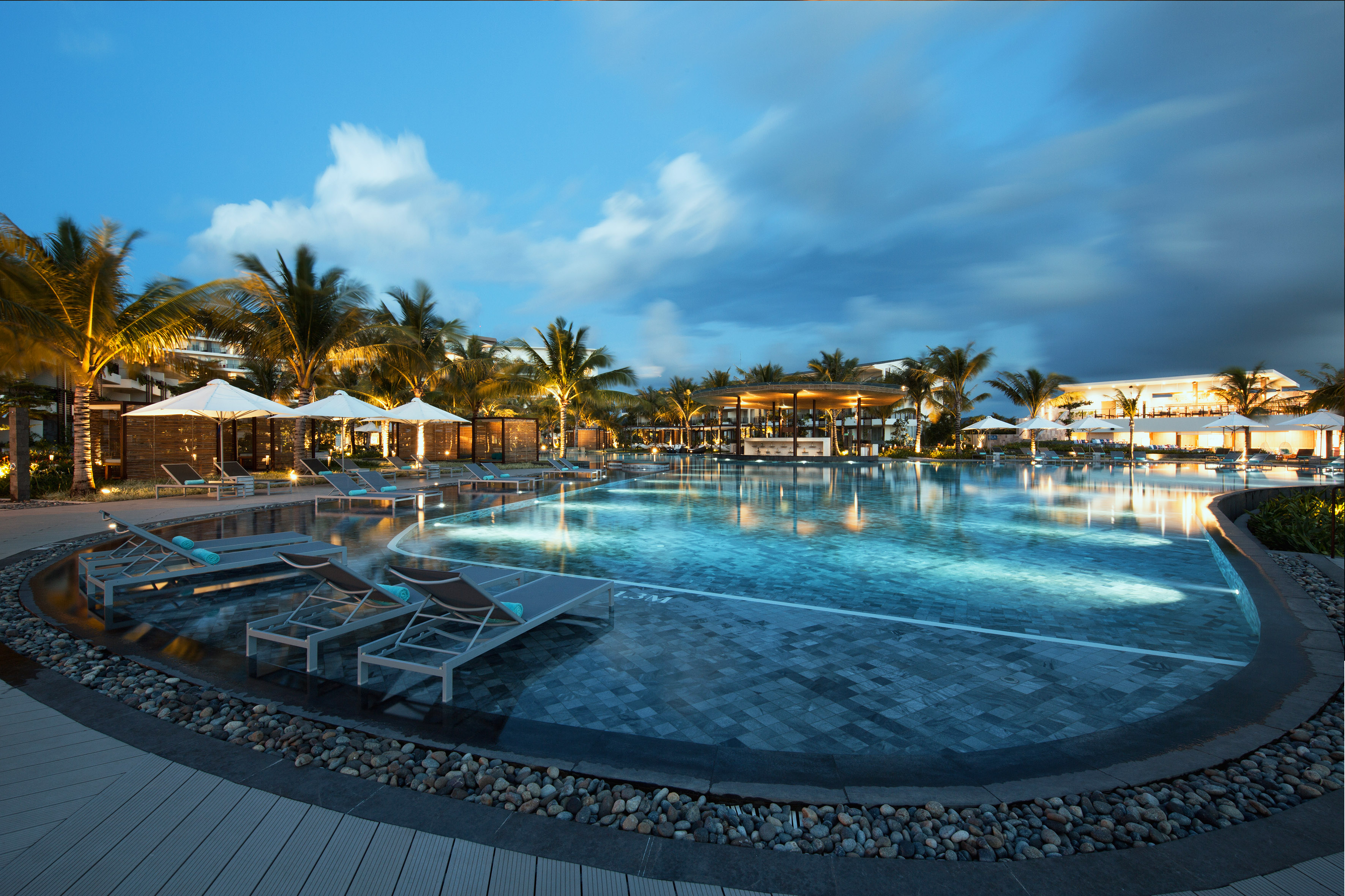 Illuminated luxury resort pool at dusk with palm trees and lounge chairs.