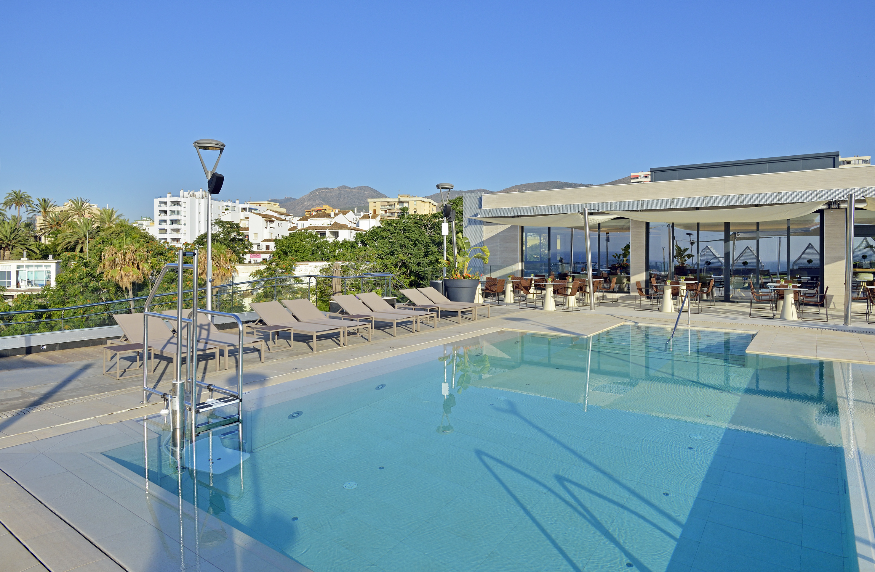 Sunny hotel rooftop pool, sun loungers, outdoor restaurant, city views.