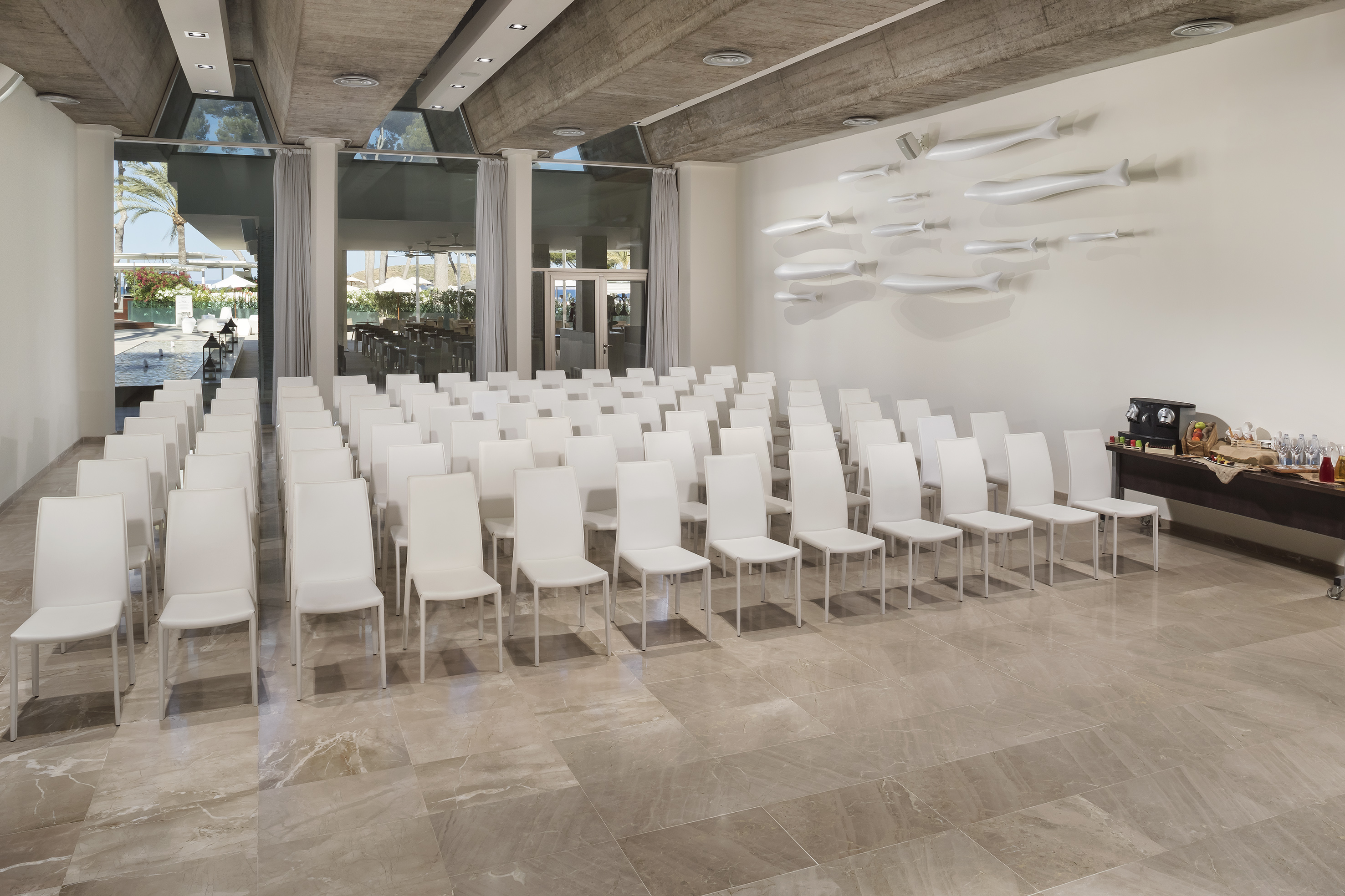 Large conference room with white chairs, refreshment table, and garden view windows.