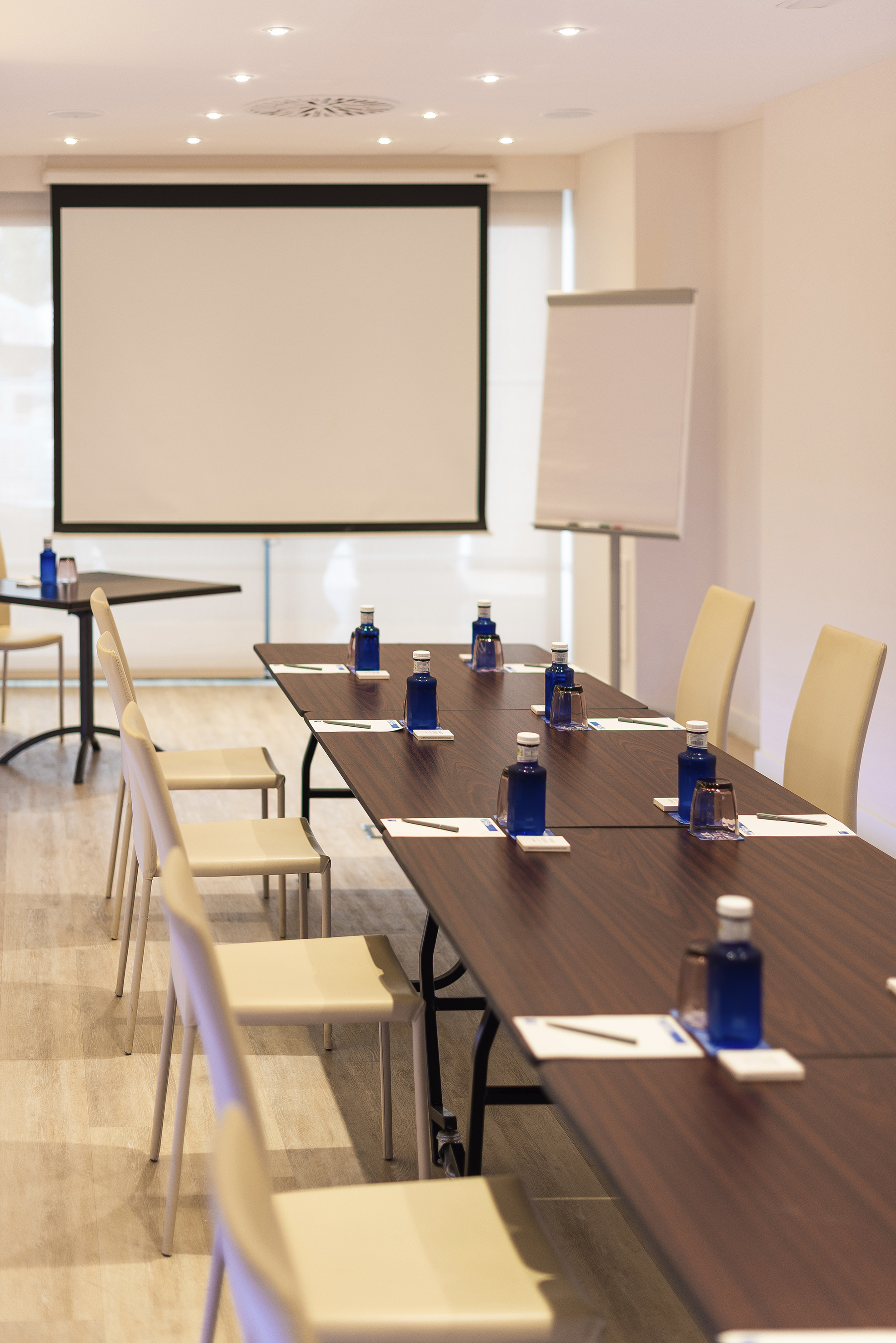 Modern conference room with tables, chairs, projector screen, and water bottles.