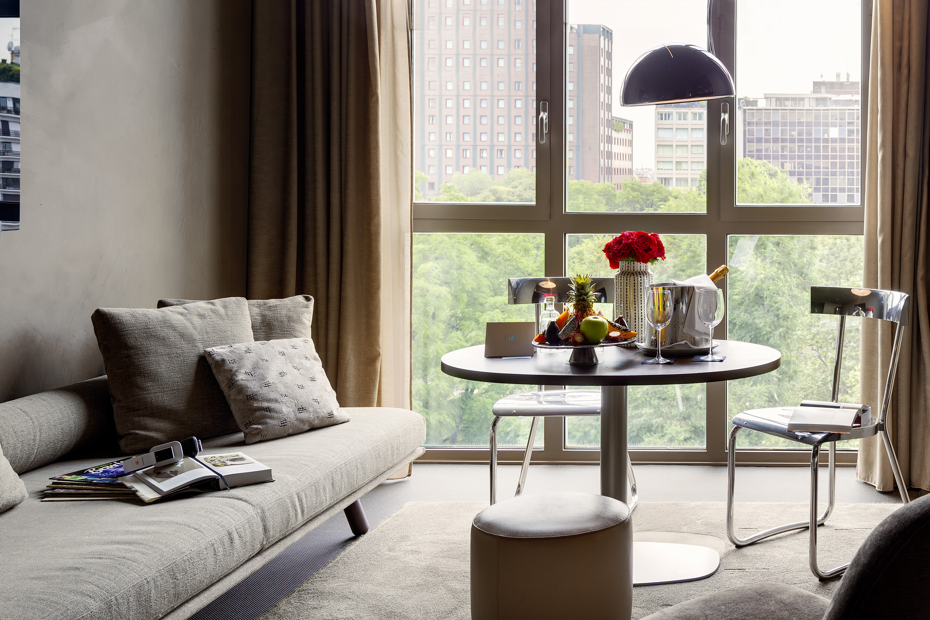 Modern hotel suite with sofa, dining table, champagne, fruit, and city view.