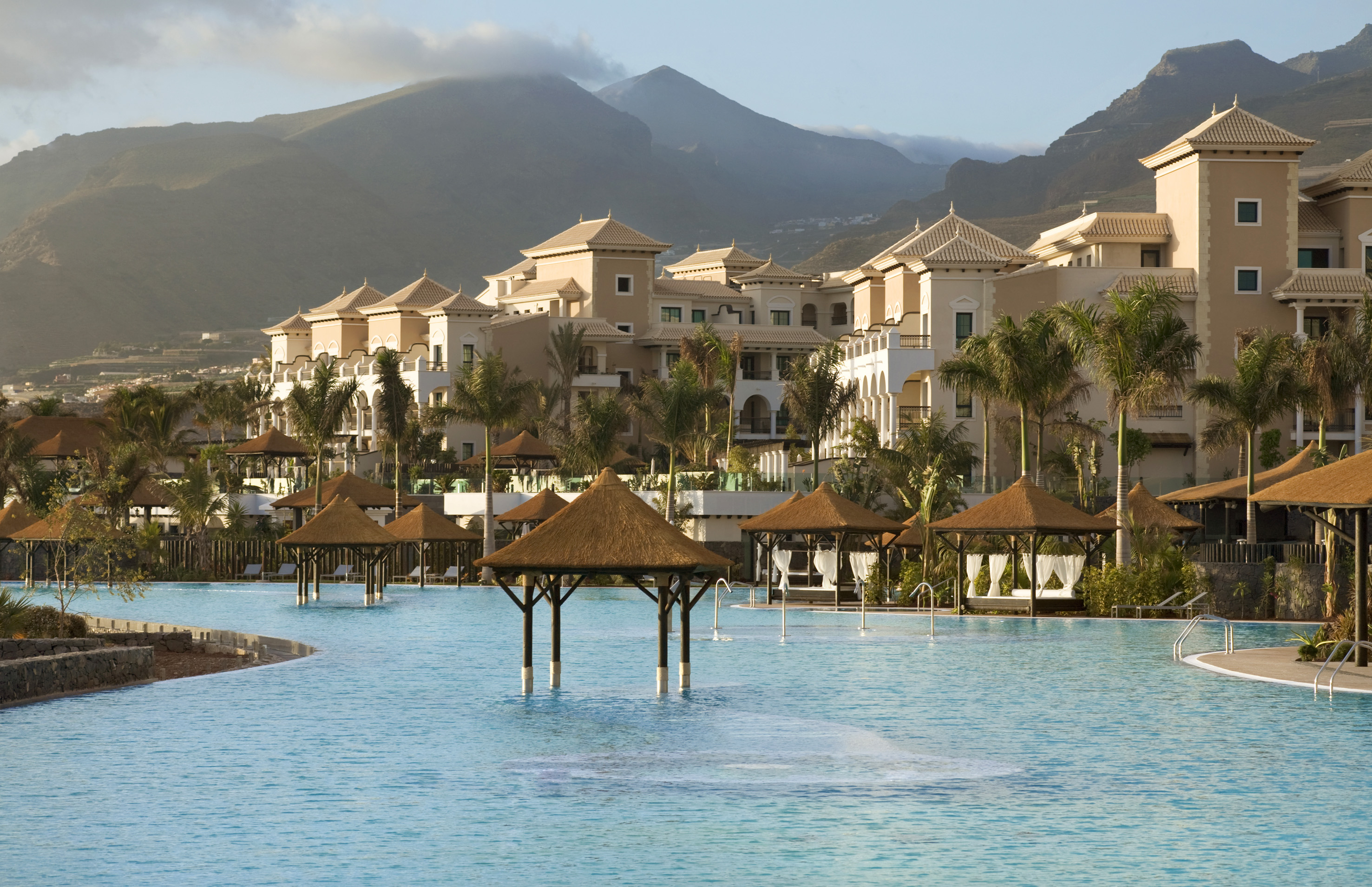 Luxury resort hotel pool with palm trees, thatched cabanas, and mountains.