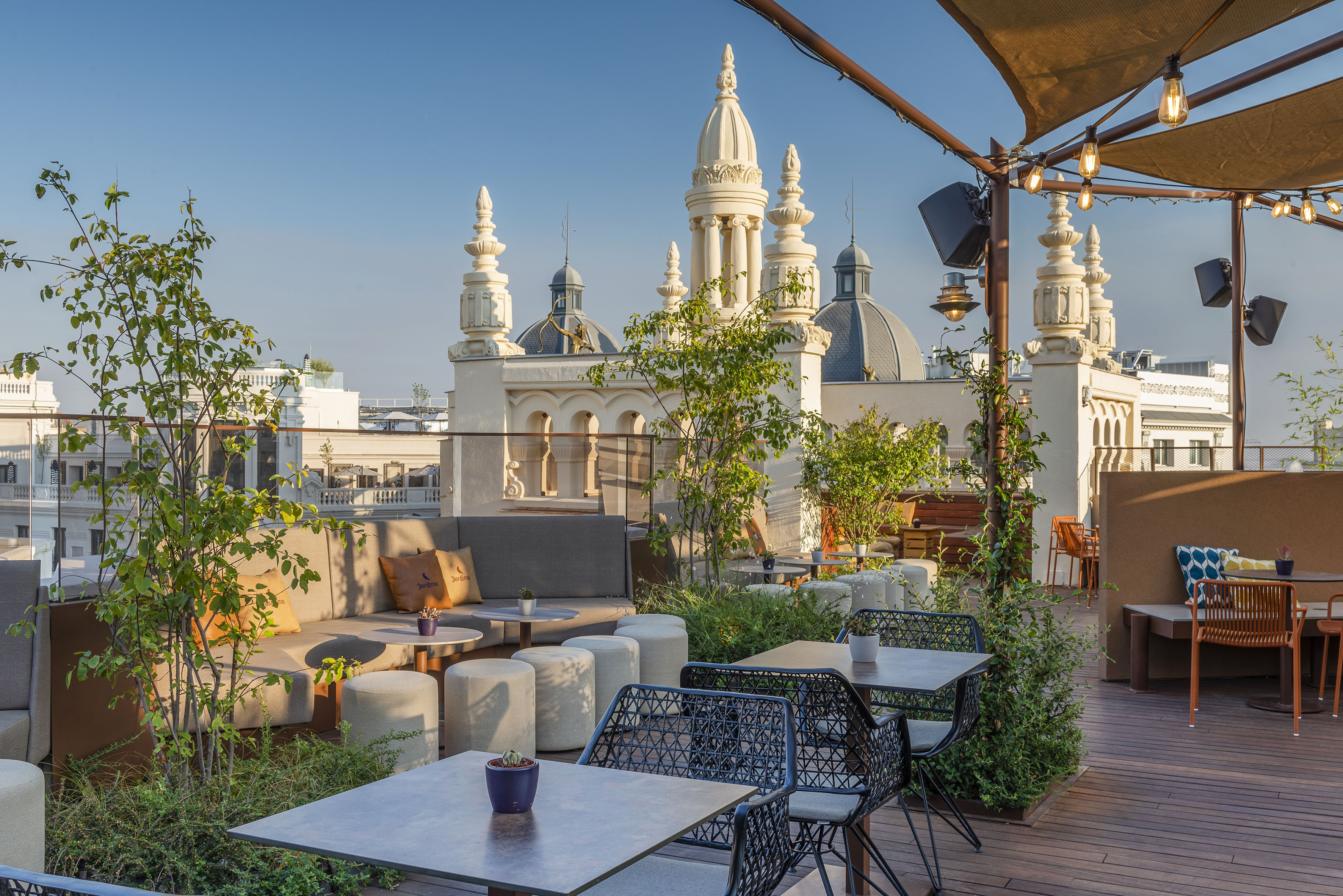 Rooftop terrace bar with outdoor seating, plants, string lights, and city spires.