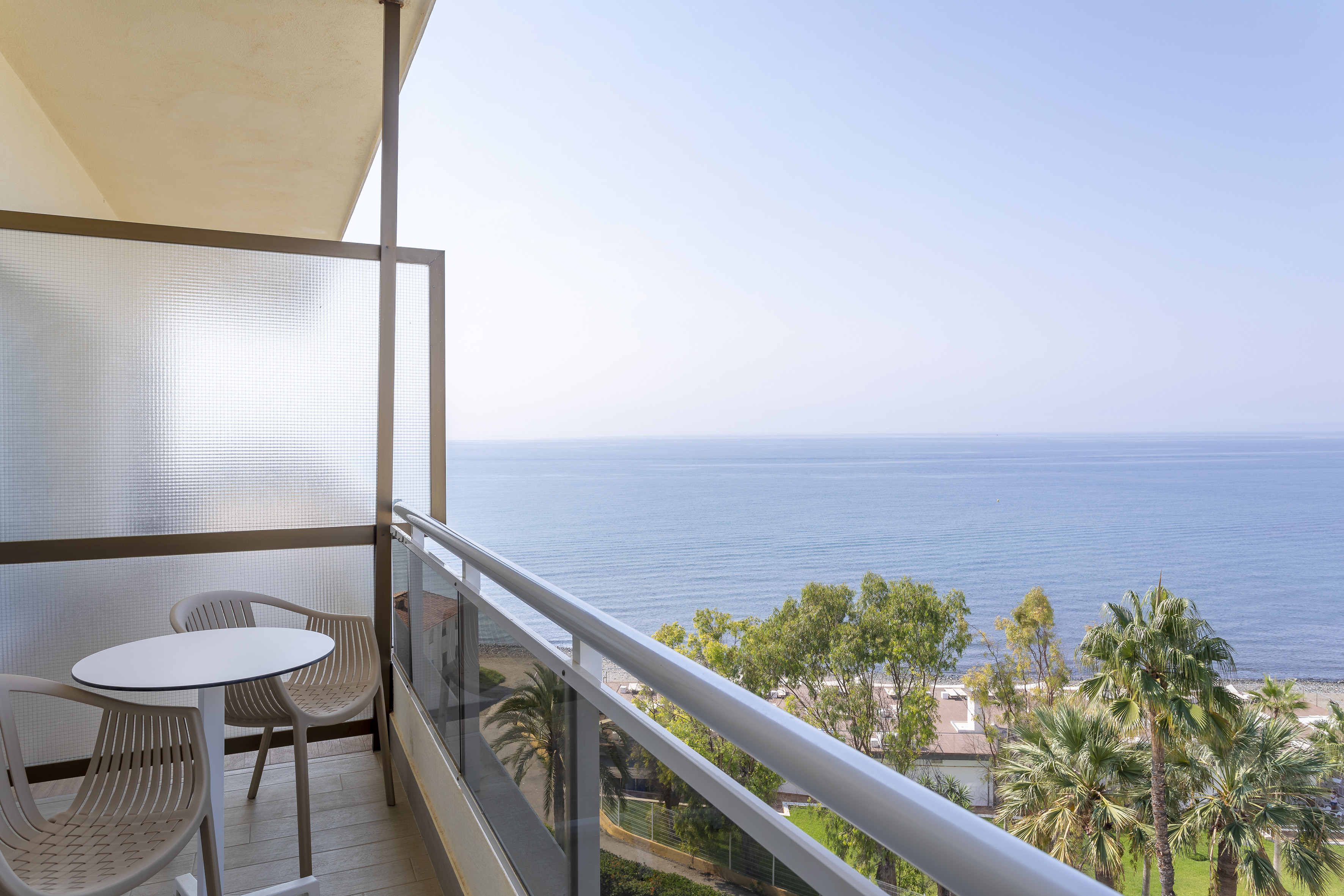 Balcony with sea view, outdoor table, chairs, palm trees, and beach.
