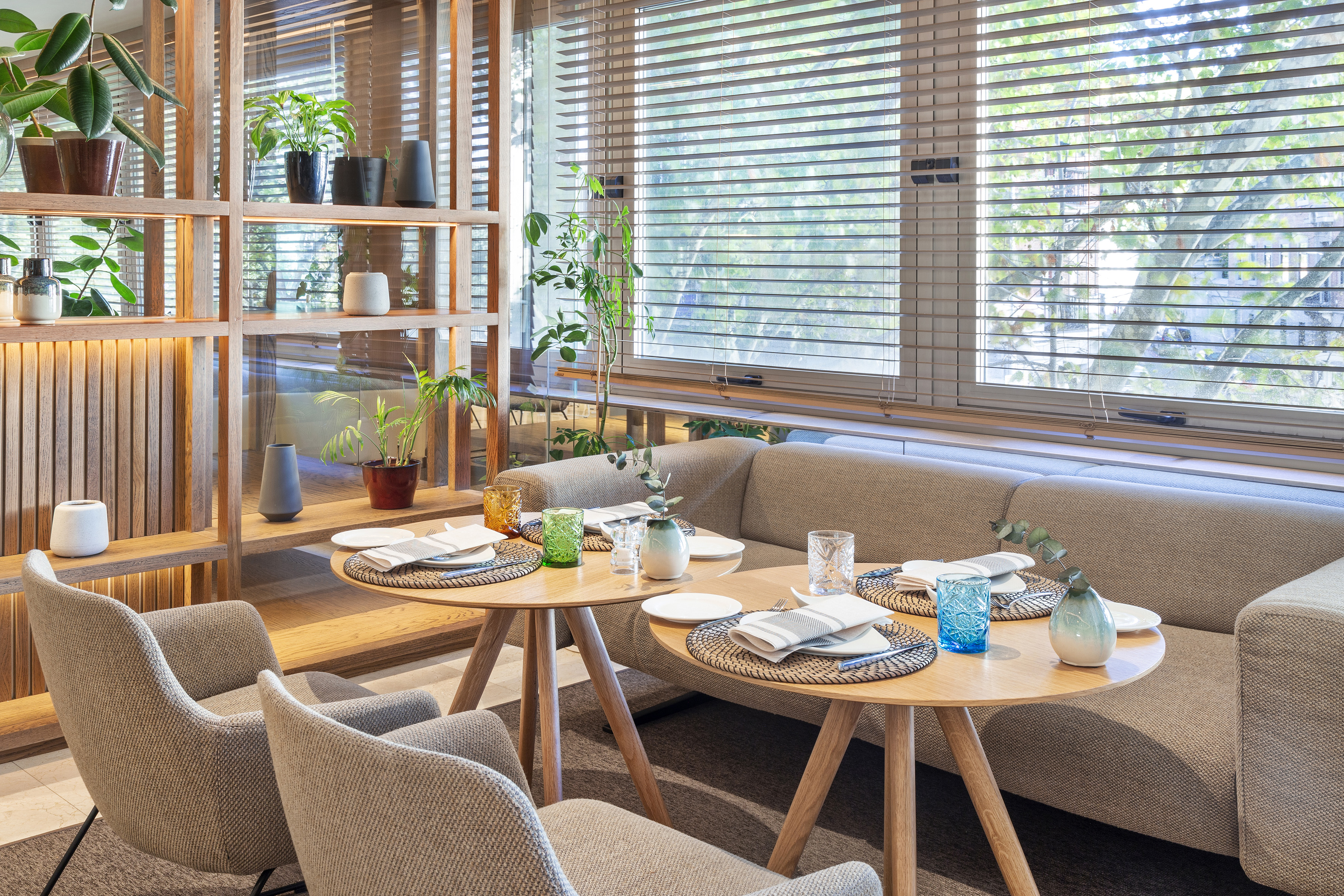 Modern restaurant dining: plants, wooden shelves, sofa seating, tables, window blinds.