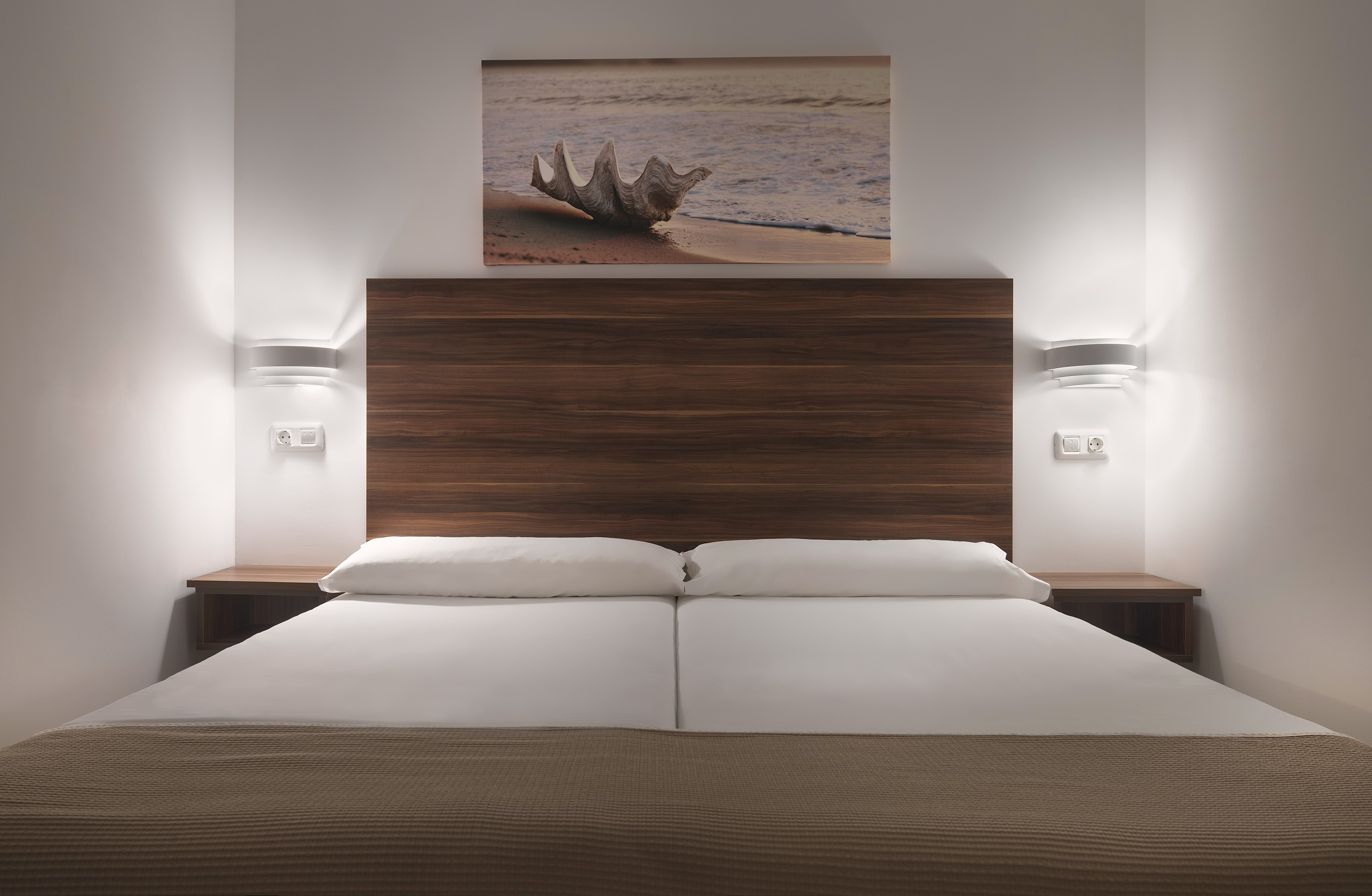 Modern hotel bedroom with wooden headboard, bed, wall lights, and beach art.