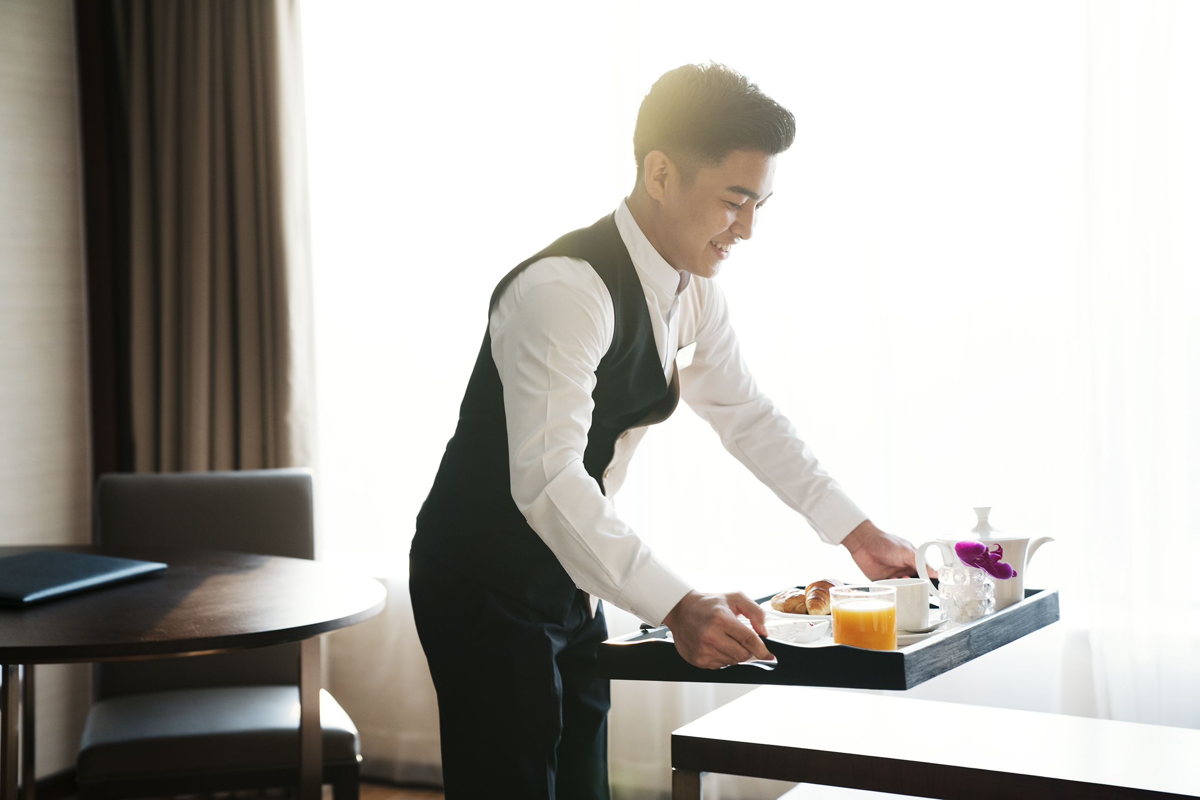 Smiling hotel staff serving breakfast tray with juice and croissants.