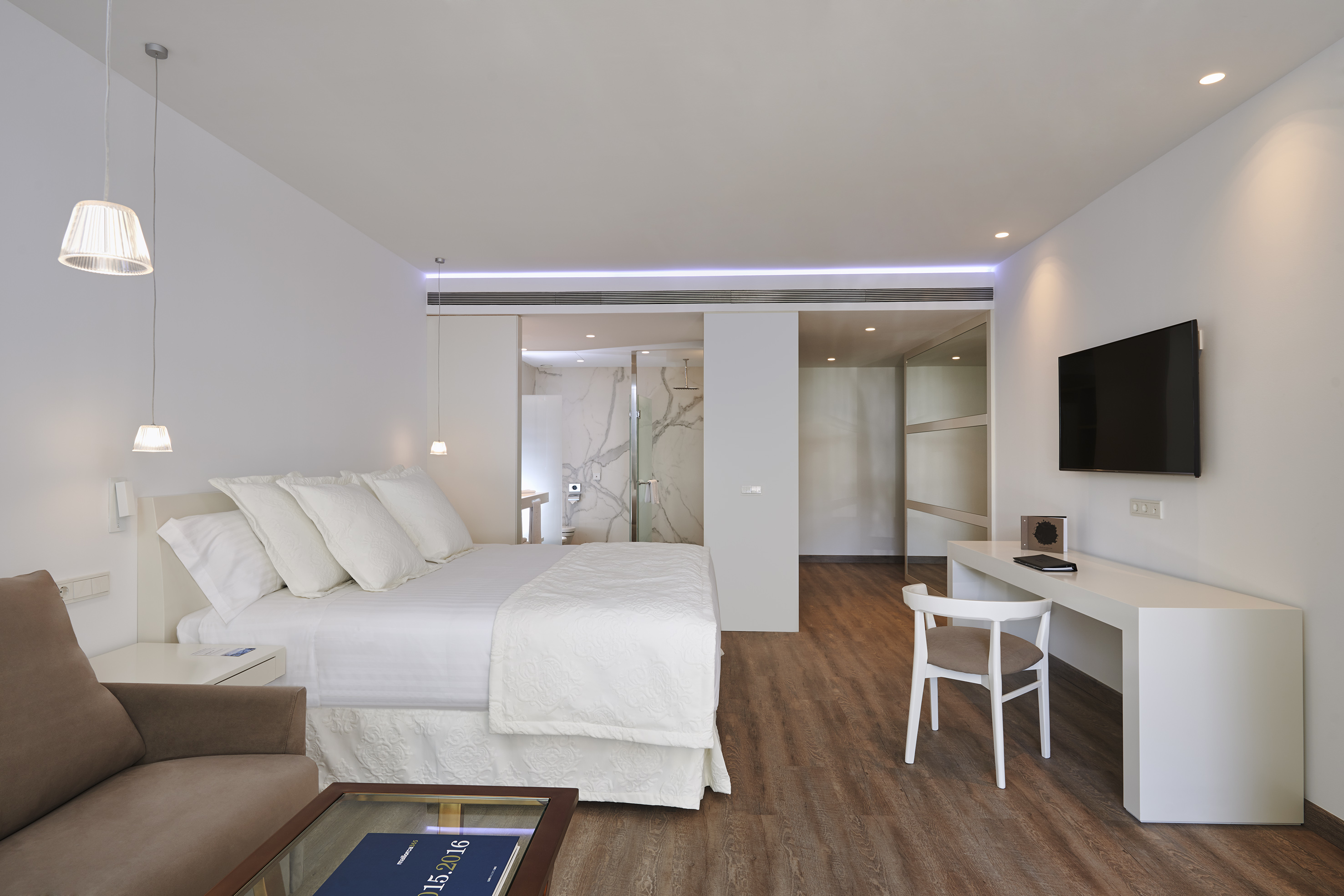Modern minimalist hotel suite with white bed, desk, sofa, TV, open bathroom.