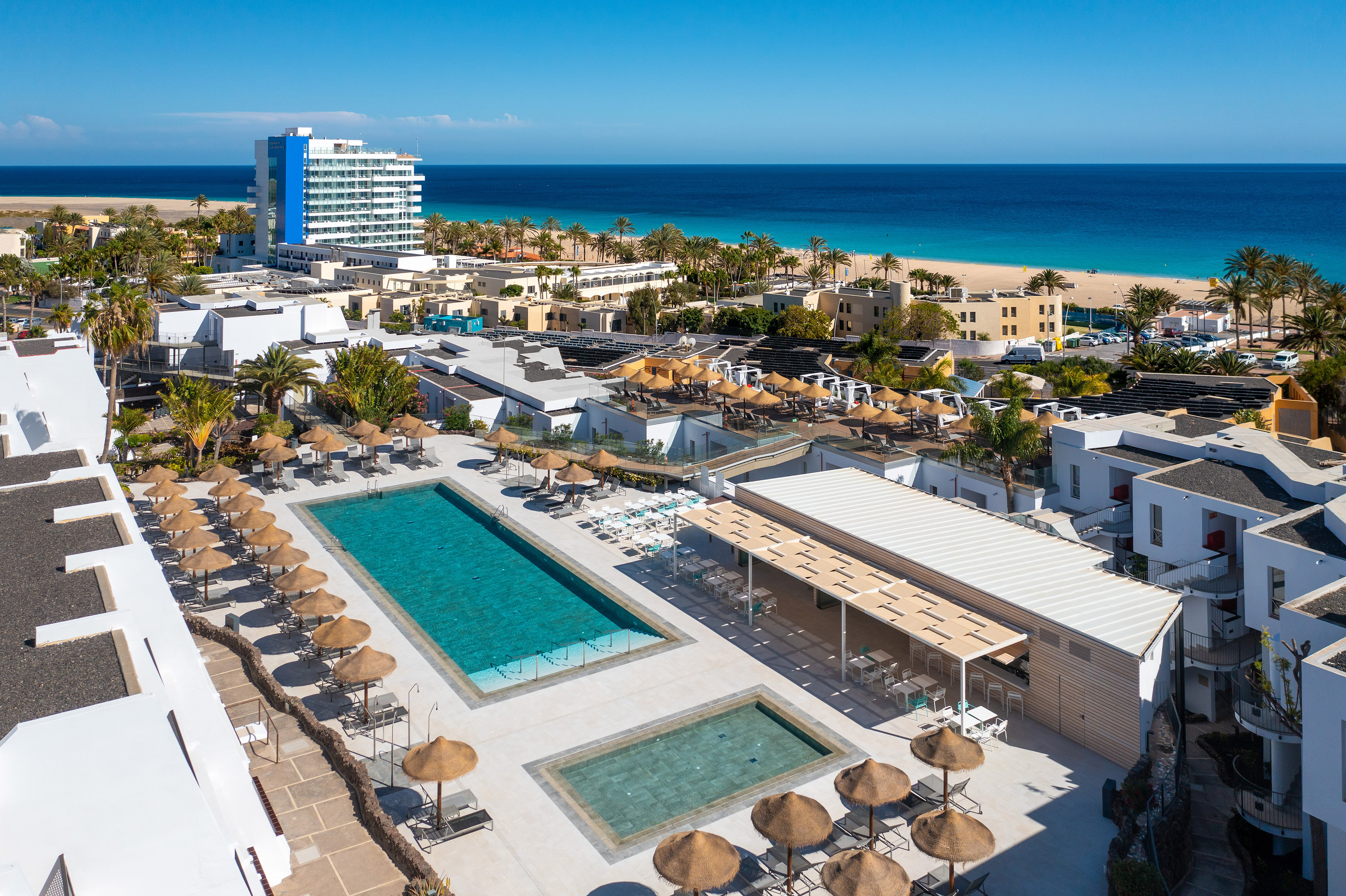 Coastal resort with swimming pools, sun loungers, palm trees, beach, and blue ocean.