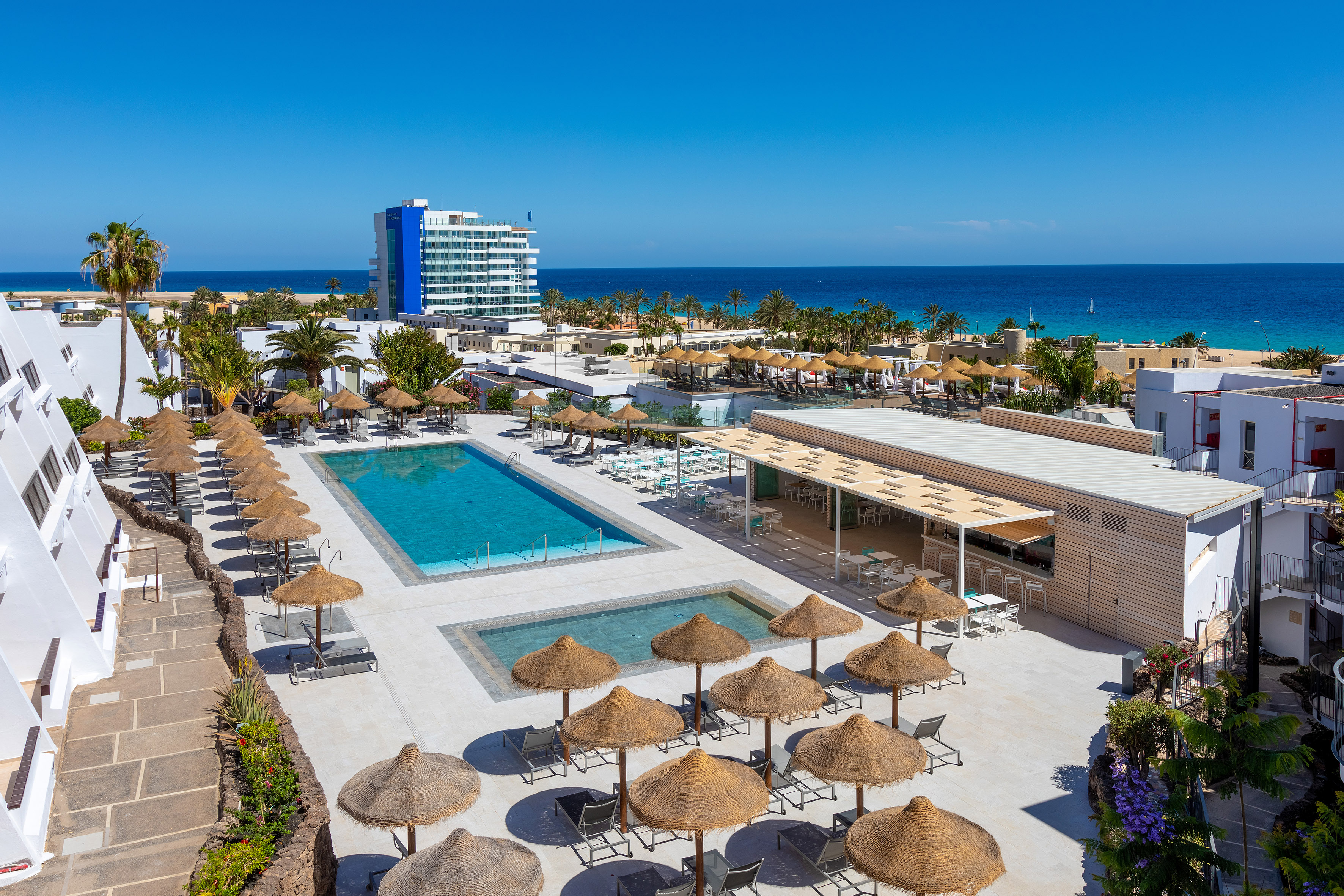 Beach resort with pools, straw umbrellas, ocean, palm trees, and hotel.