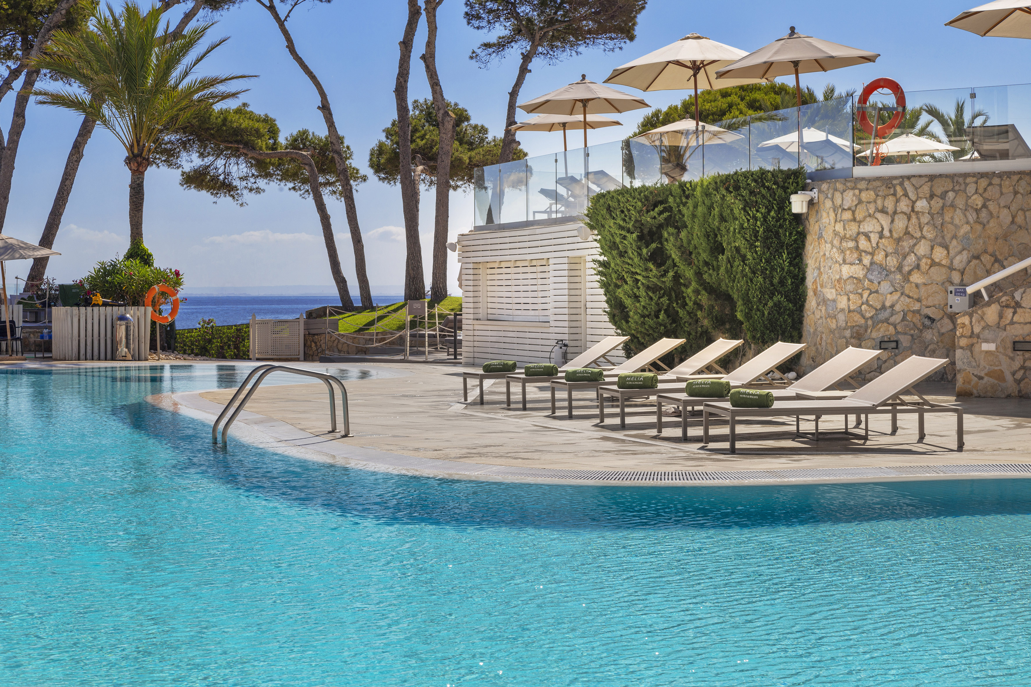 Resort swimming pool with lounge chairs, parasols, palm trees, and ocean view.