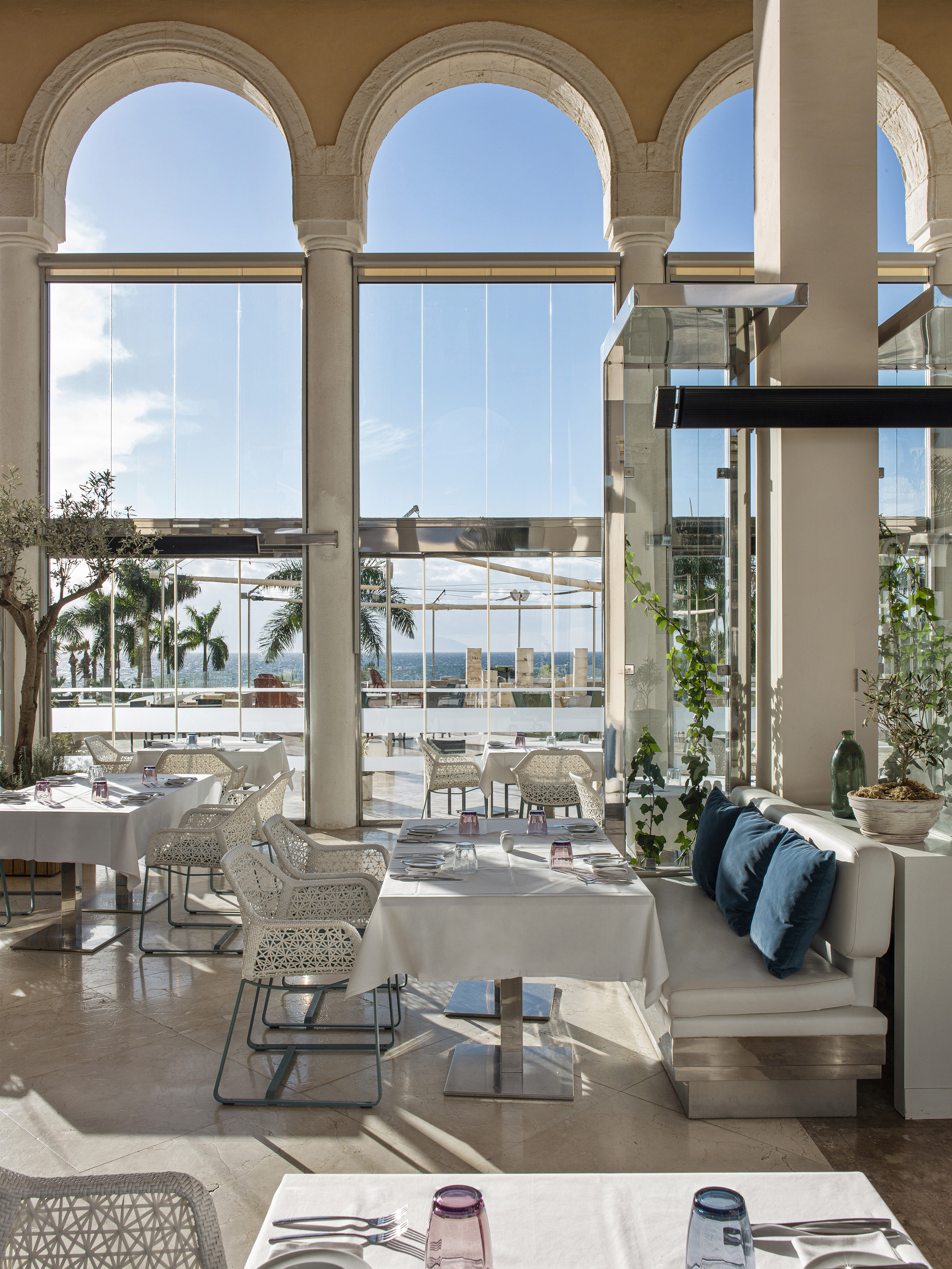 Sunny restaurant with panoramic sea view through arched windows, white tables.
