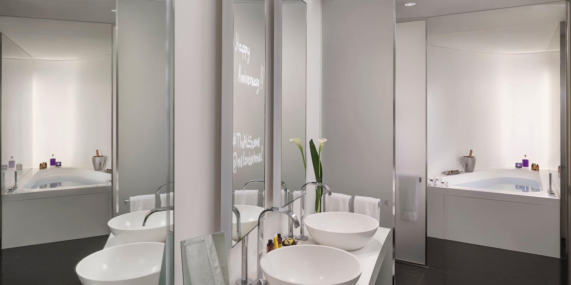 Modern hotel bathroom: dual sinks, tub, Happy Anniversary on mirror.