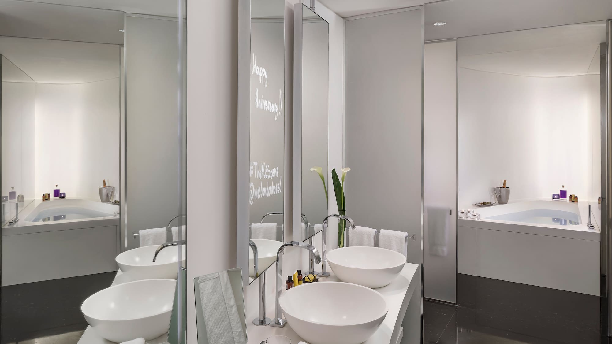 Modern hotel bathroom: dual sinks, tub, Happy Anniversary on mirror.