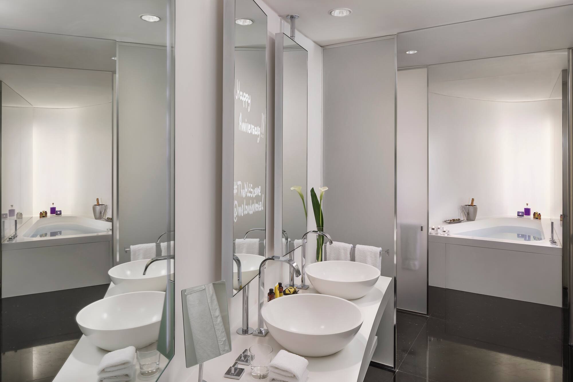 Modern hotel bathroom: dual sinks, tub, Happy Anniversary on mirror.