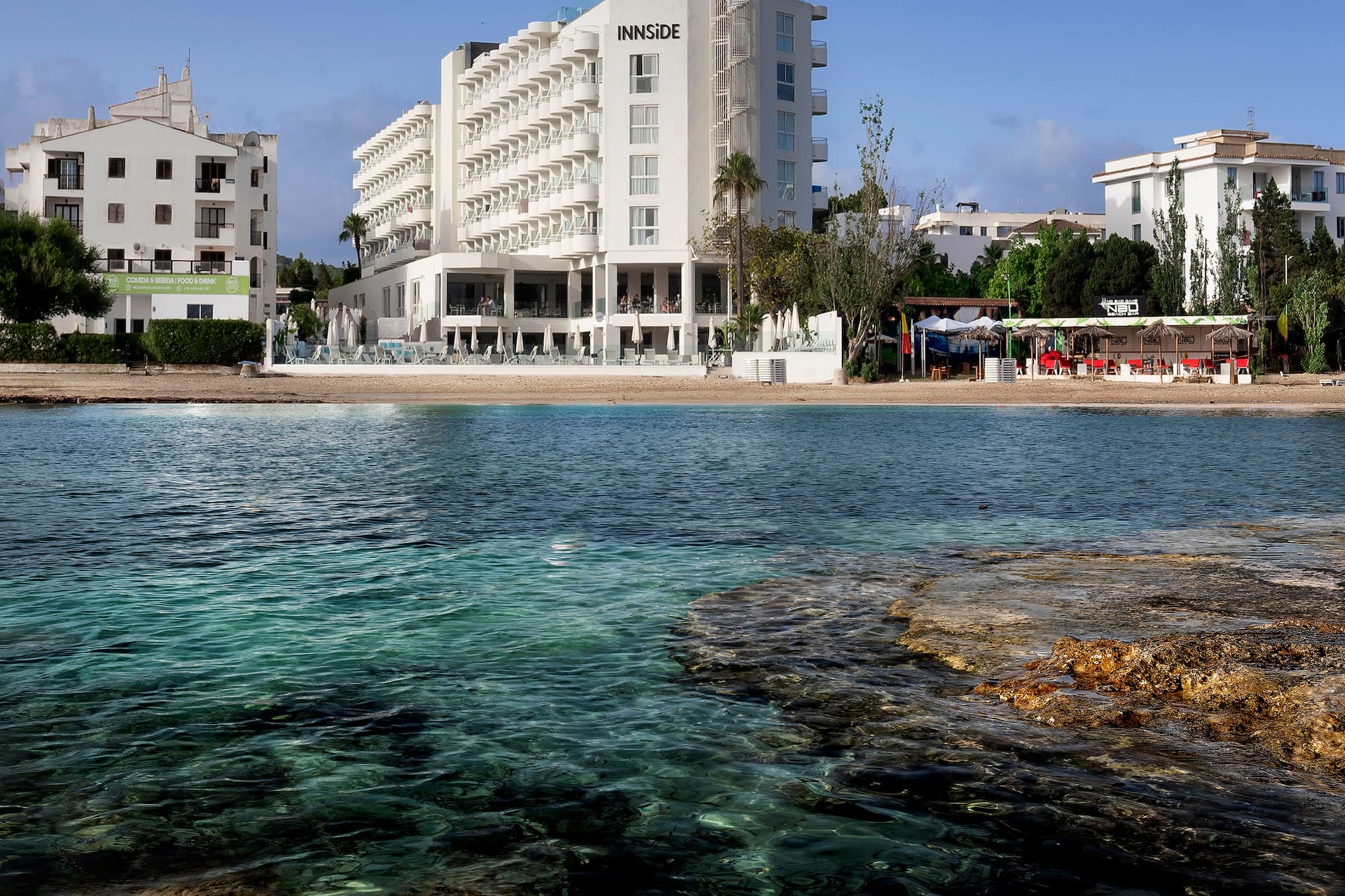 Innside hotel on a sandy beach with clear turquoise sea and rocky foreground.