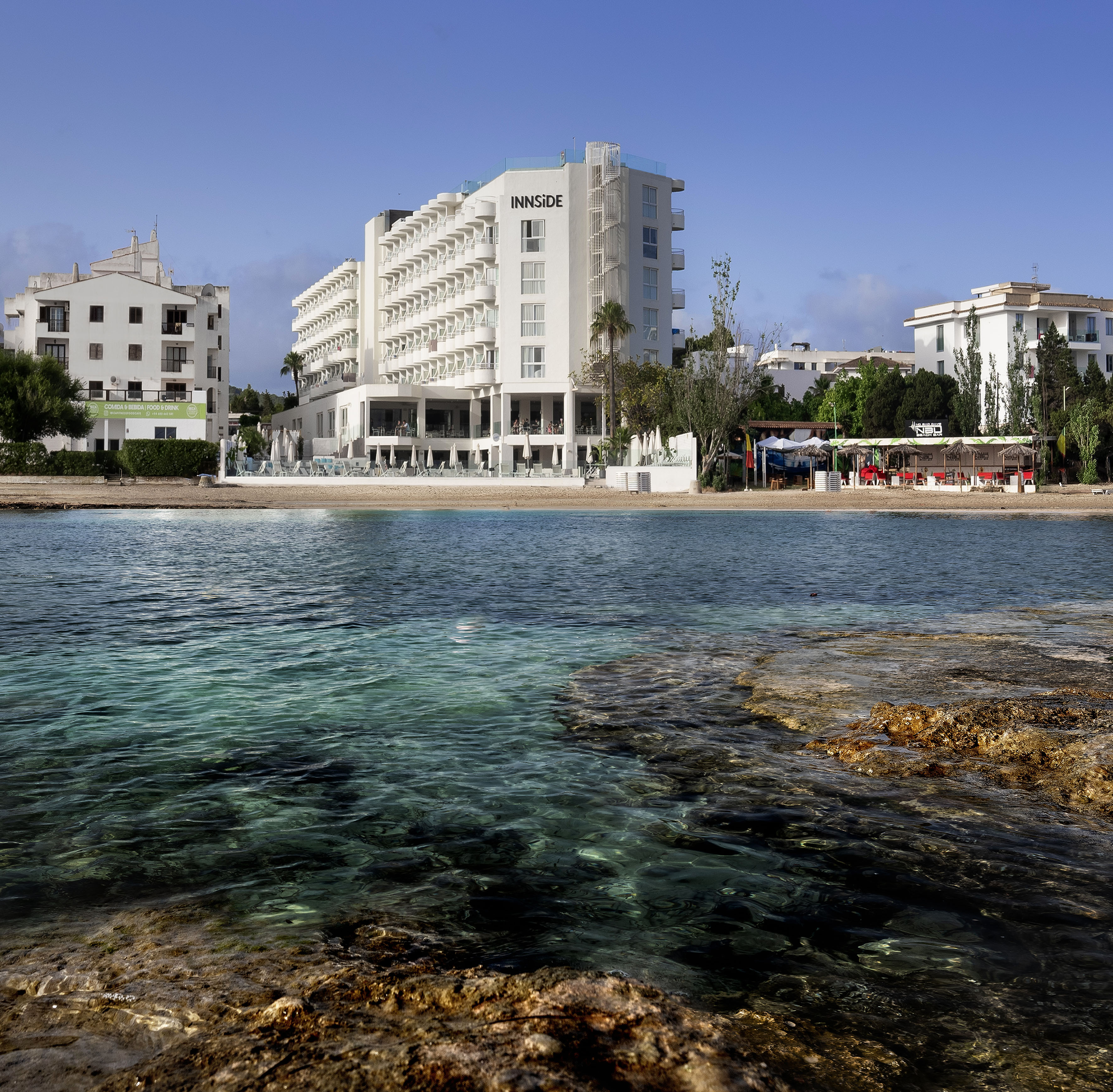 Innside hotel on a sandy beach with clear turquoise sea and rocky foreground.