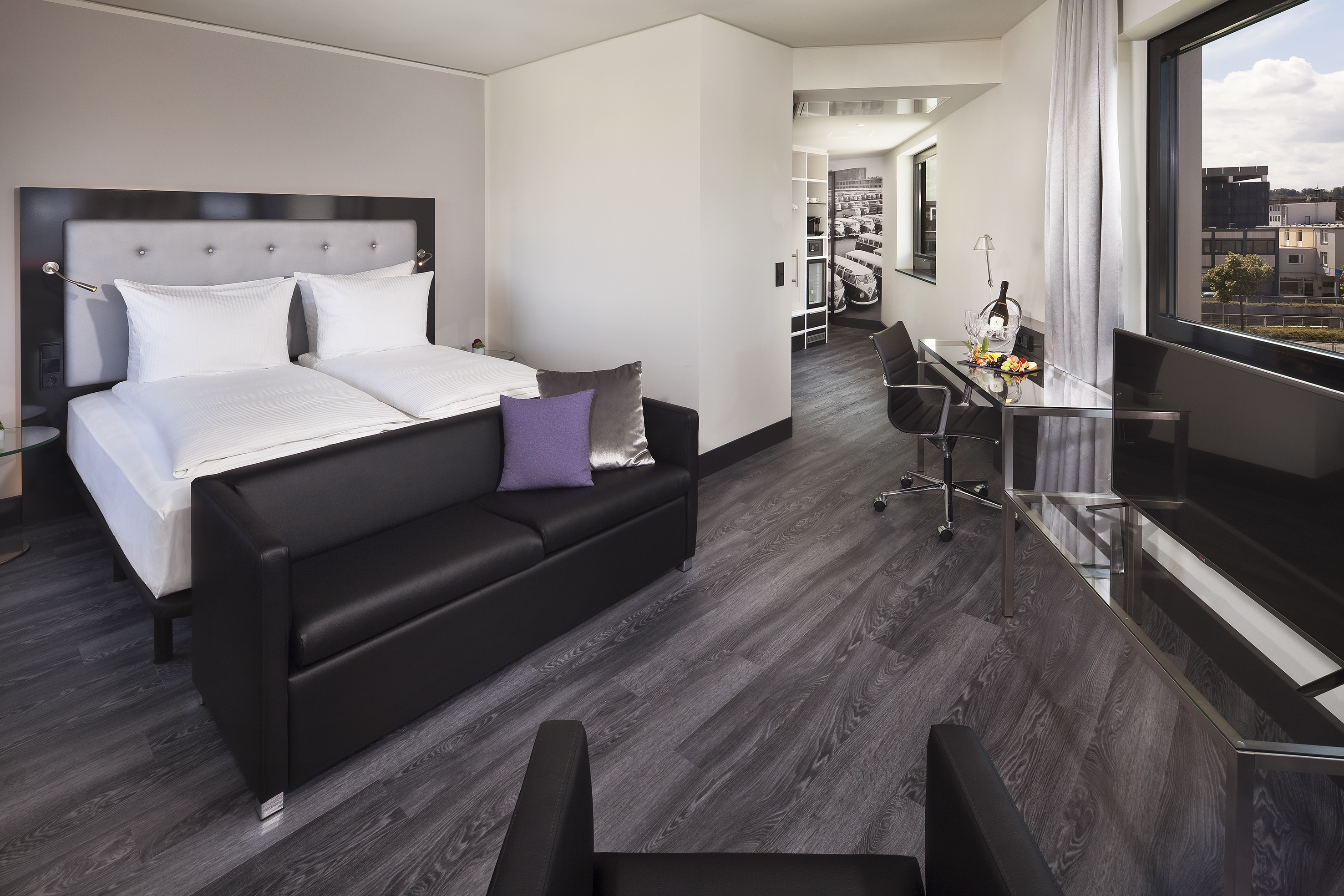 Spacious modern hotel suite with bed, black sofa, desk, and city view.