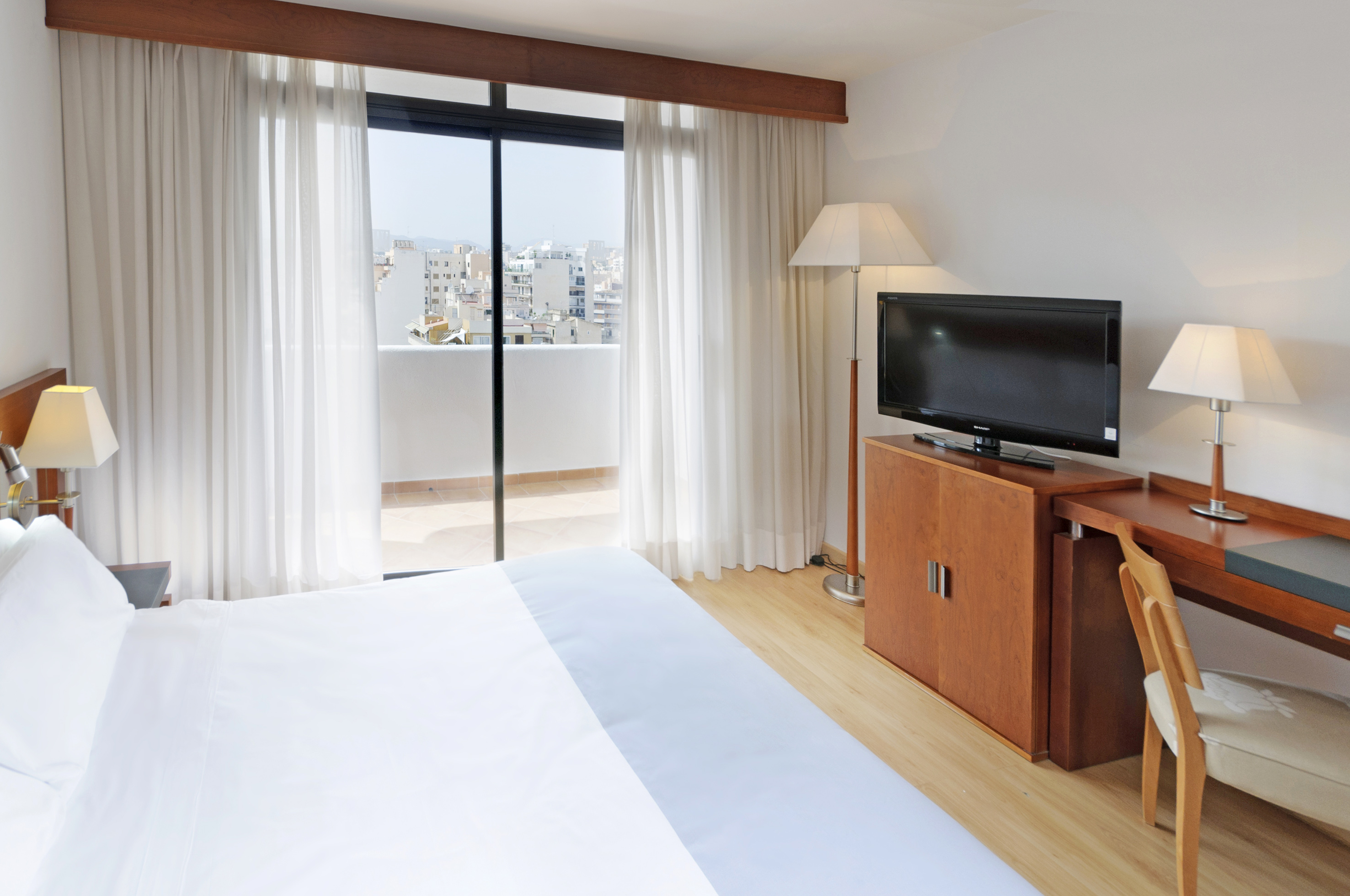 Modern hotel room with balcony city view, king bed, TV, and desk.
