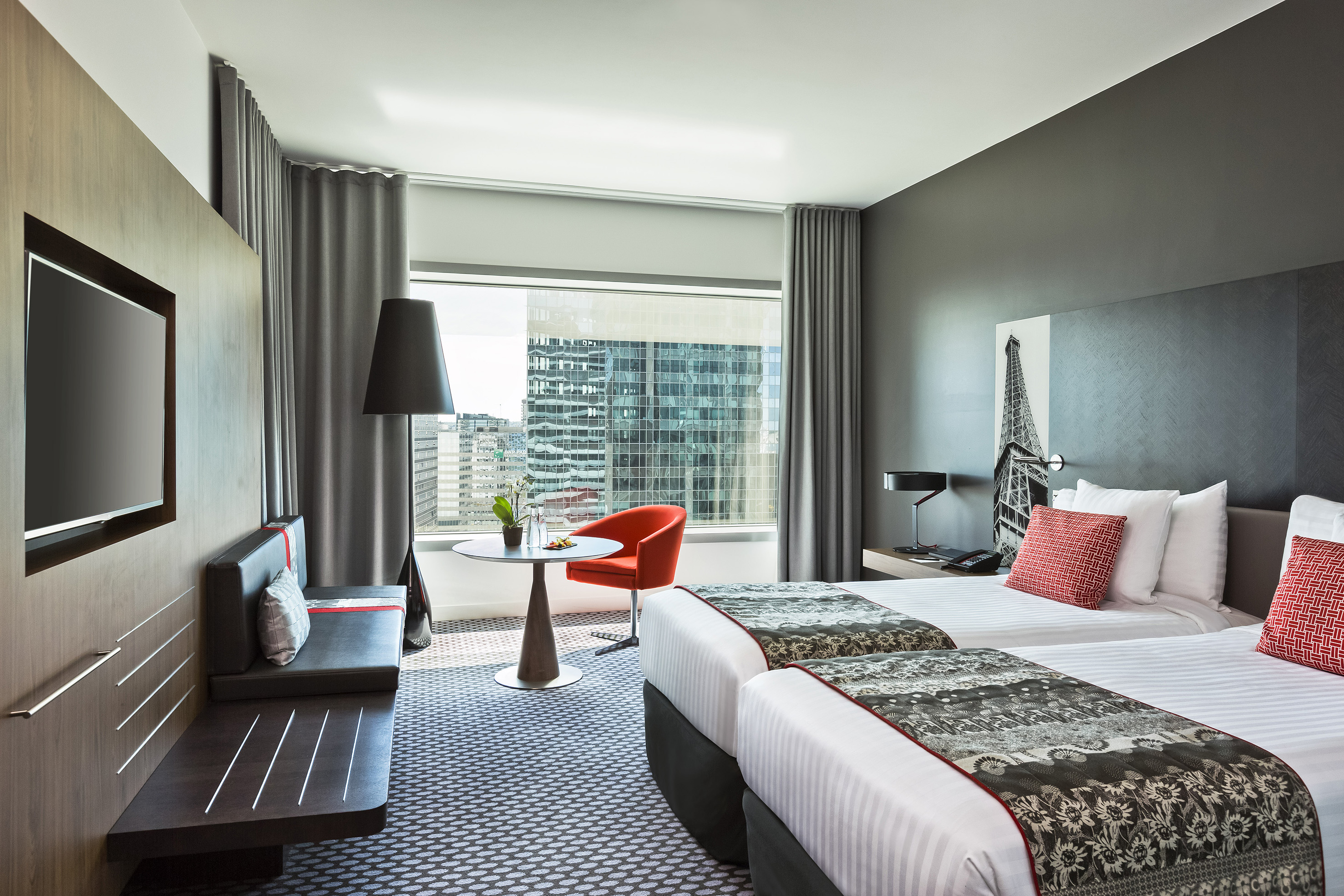 Modern hotel room with twin beds, city view, and red accents.
