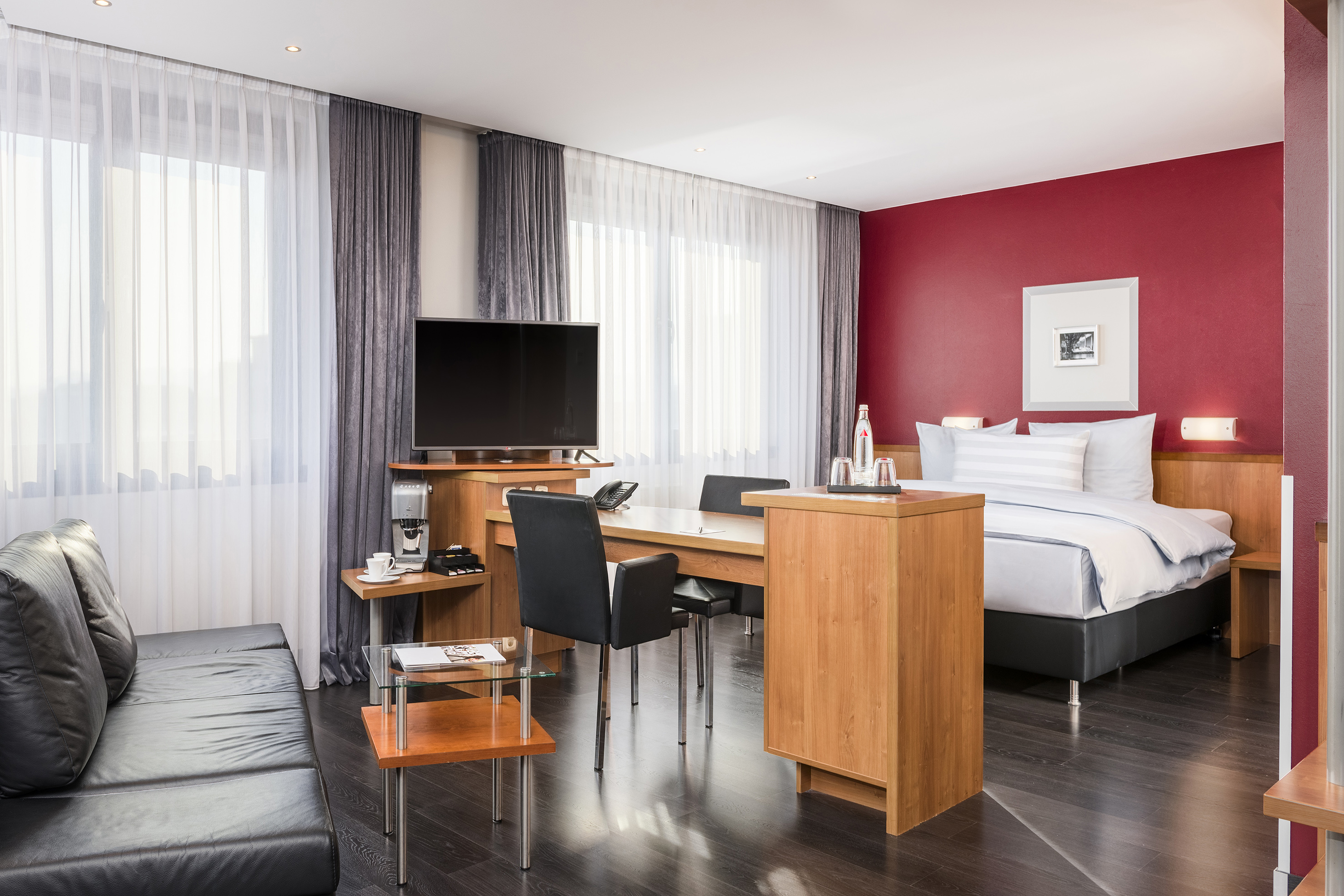 Modern hotel suite with bed, black sofa, work desk, and red accent wall.