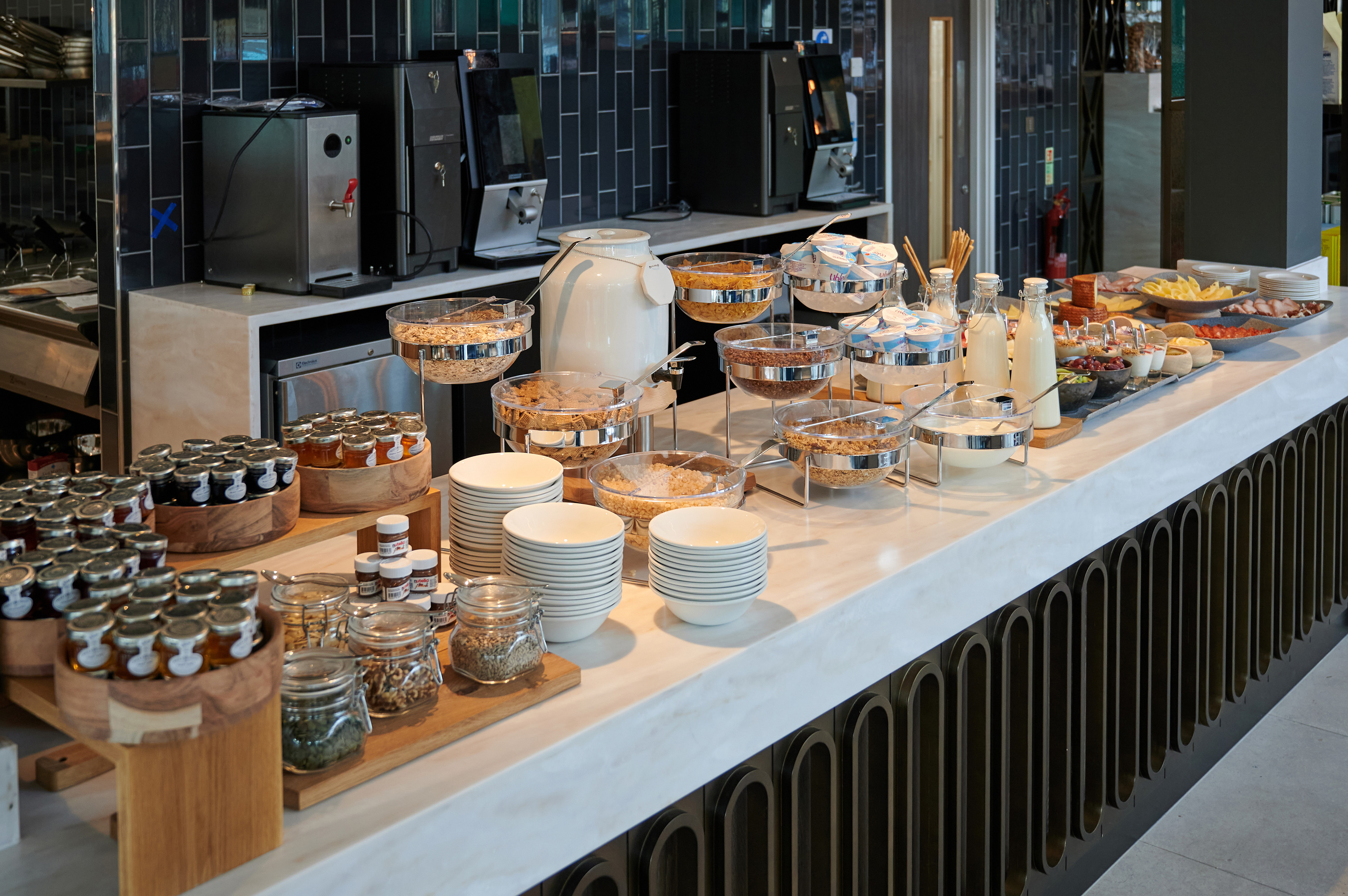 Hotel breakfast buffet with cereals, yogurt, milk, jams, and coffee machines.